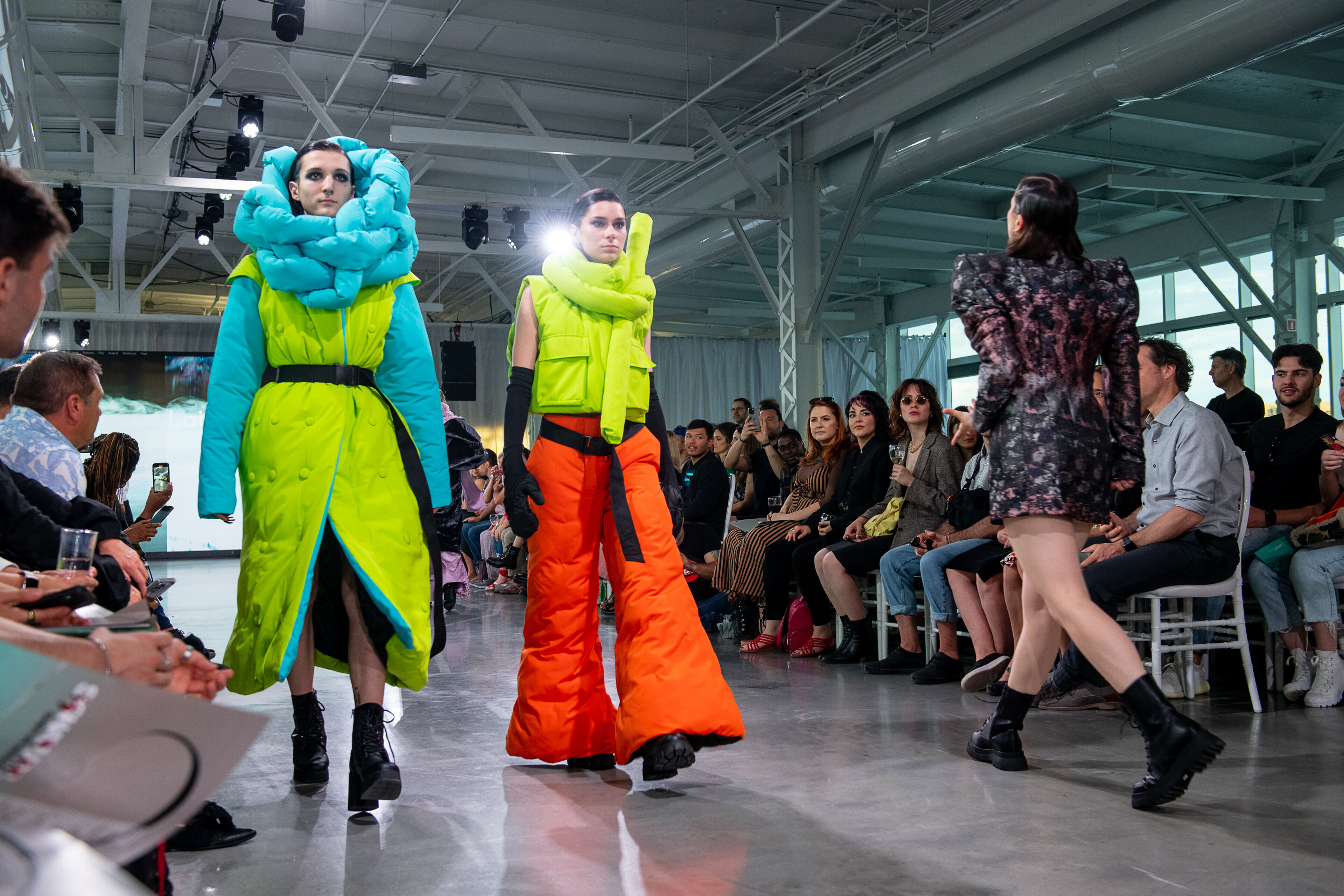 Models display bold avant-garde outfits on a runway, with an audience of spectators in the background.