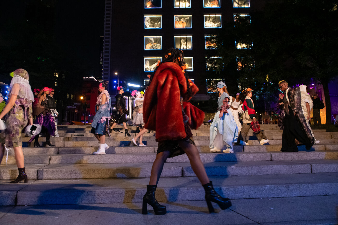 Diverse group of people in colorful costumes parading on urban steps at night, illuminated by building lights and vibrant city life.