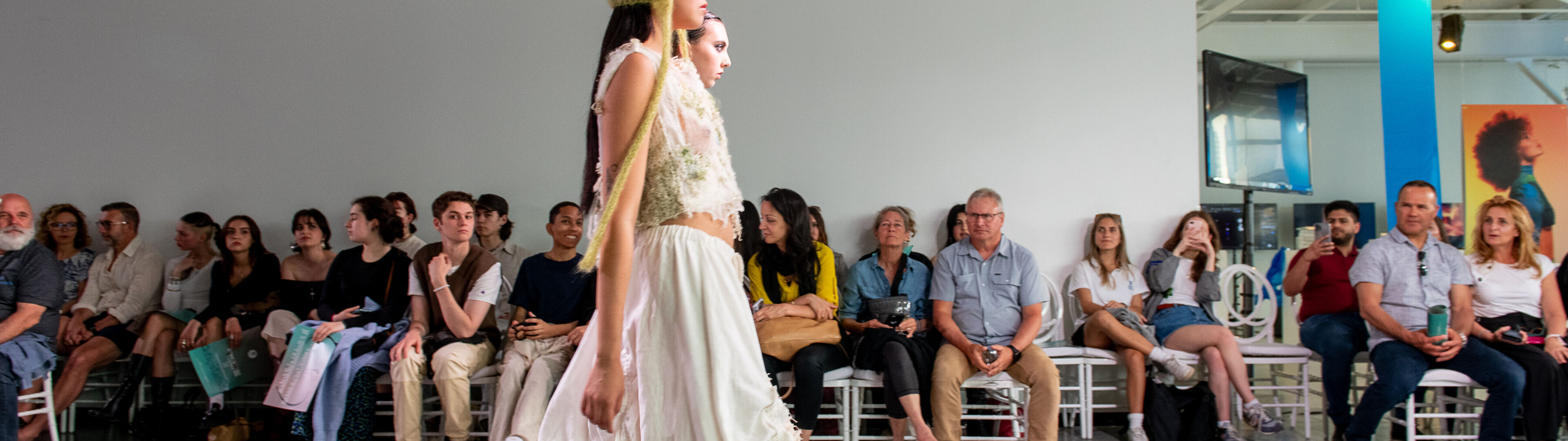 A model walks down the runway in an elegant dress with a unique headdress, audience members watching intently.