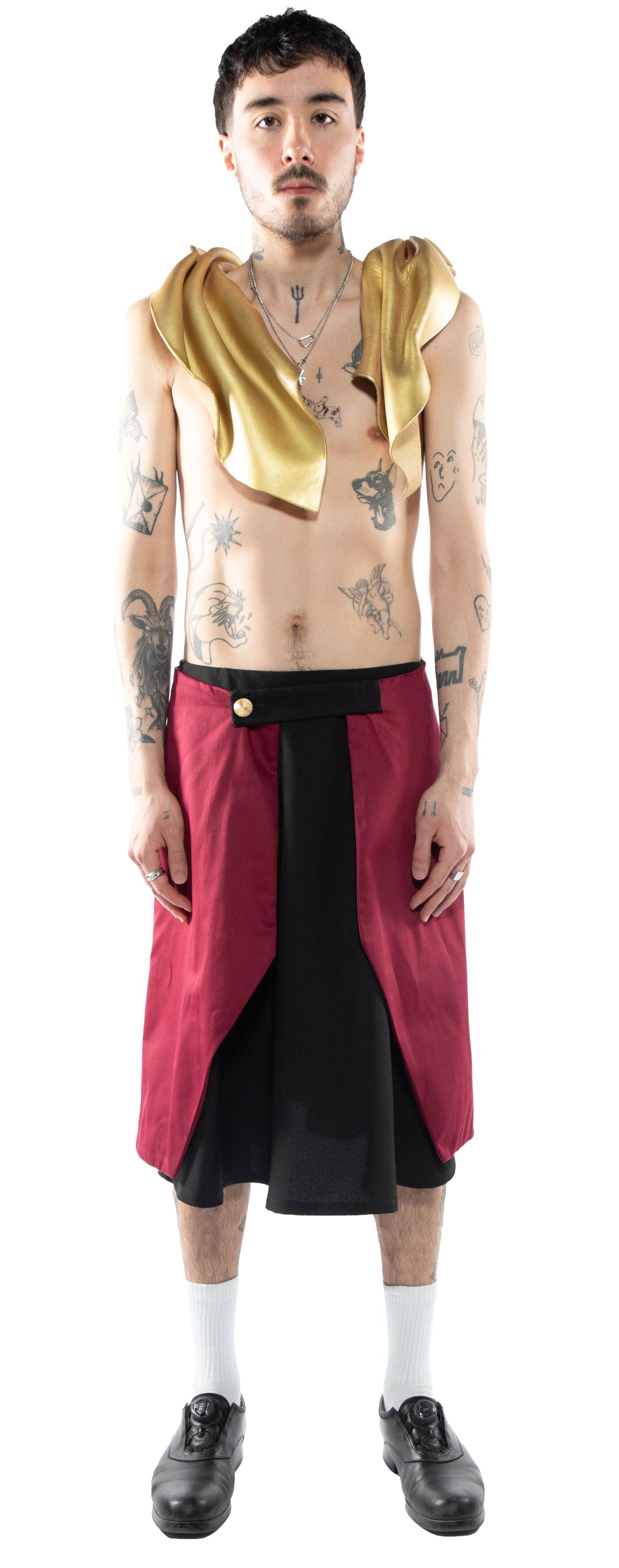 A tattooed man with a gold draped around his neck wears a burgundy and black kilt-like skirt, white socks, and black shoes.