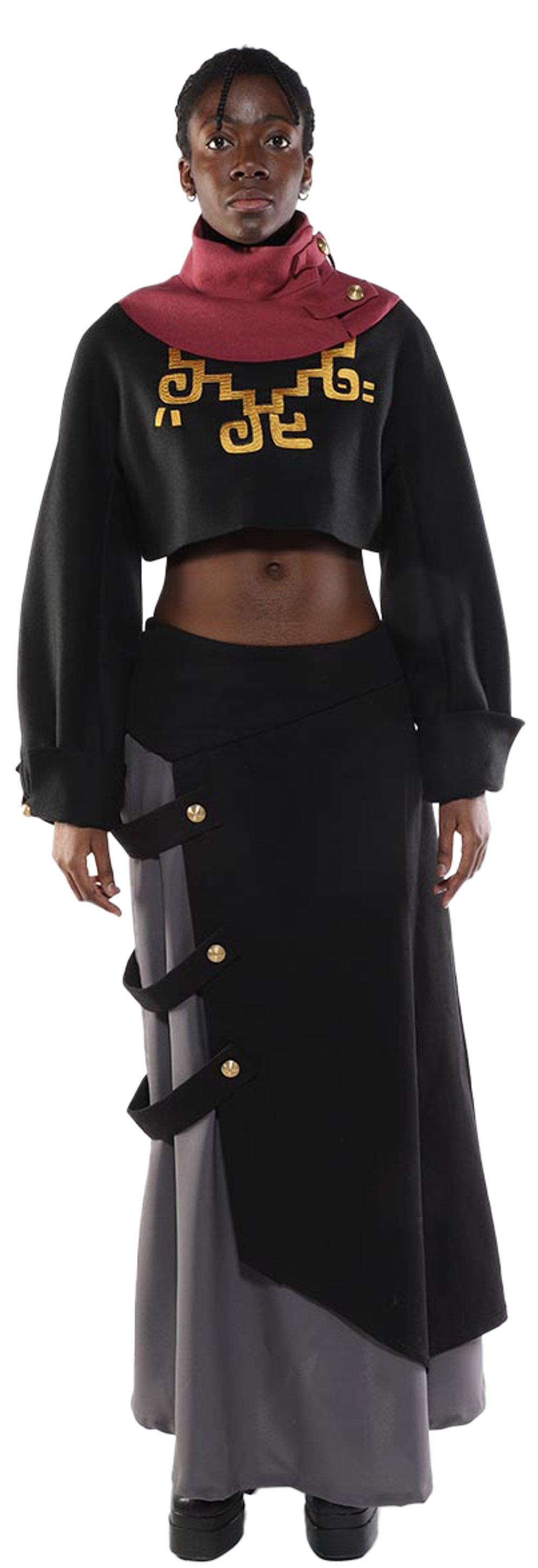 A dark-skinned model with short hair is standing against a white background wearing a black cropped sweater with a burgundy neck warmer featuring gold embroidery, and a black and gray asymmetrical midi skirt with gold buttons.