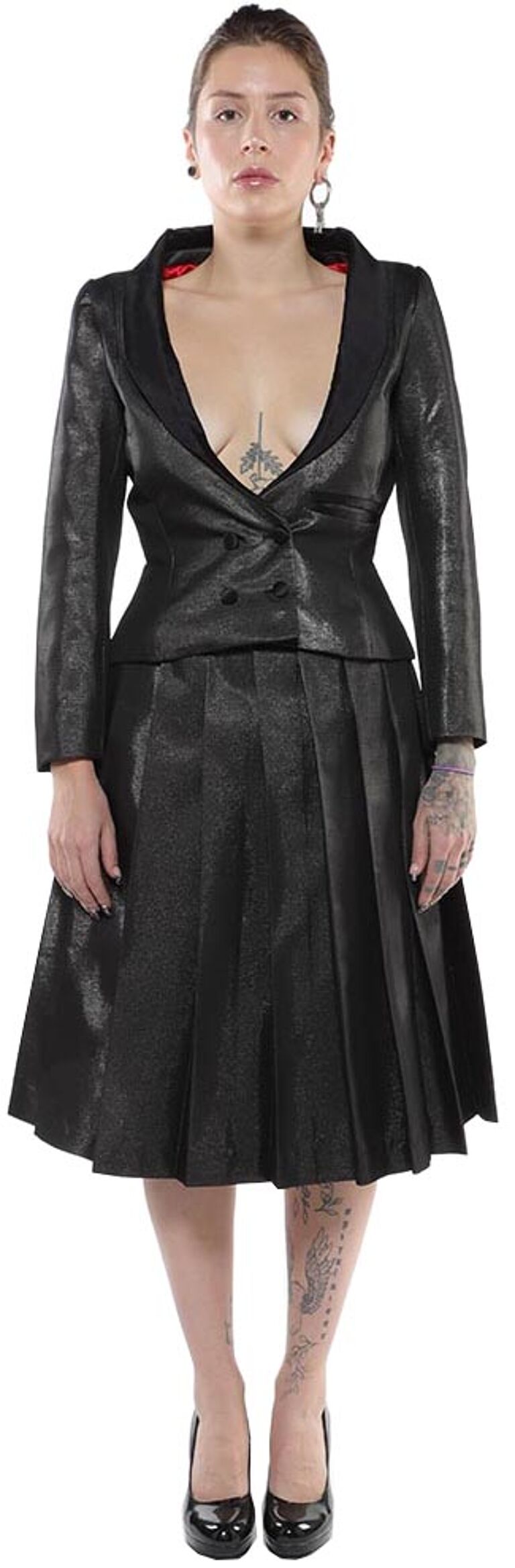 A woman with dark hair and tattoos stands wearing a black pleated skirt suit and black high heels.