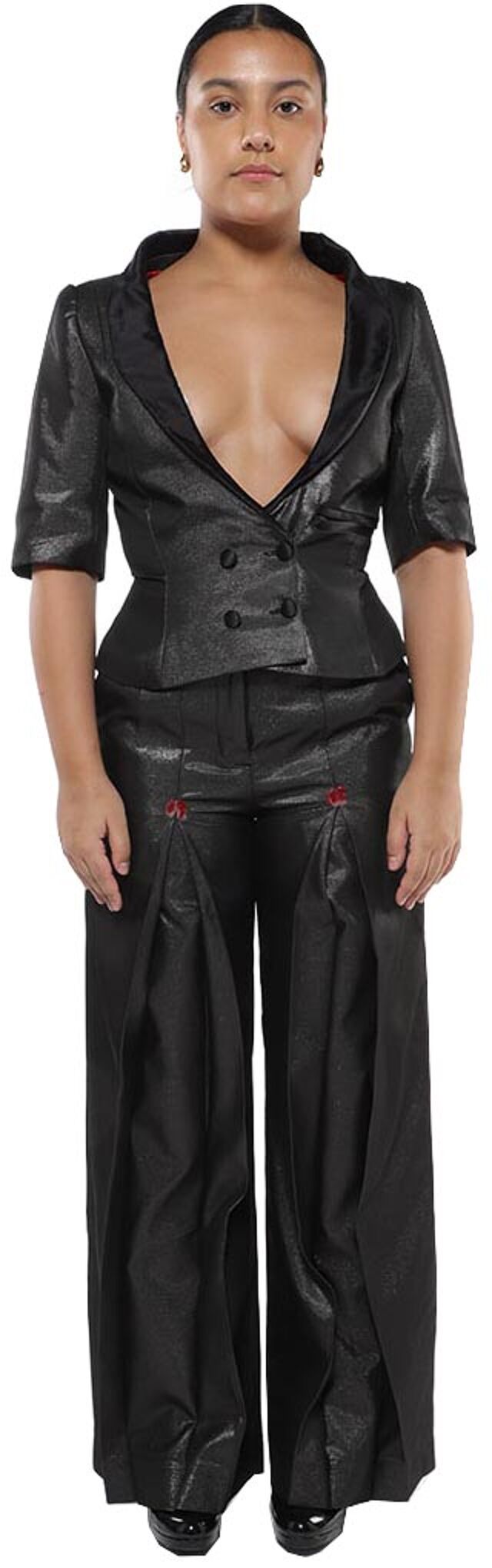 A woman with dark hair and tan skin wears a black, double-breasted, short-sleeved jacket and matching wide-legged pants with red accents, standing against a white background.
