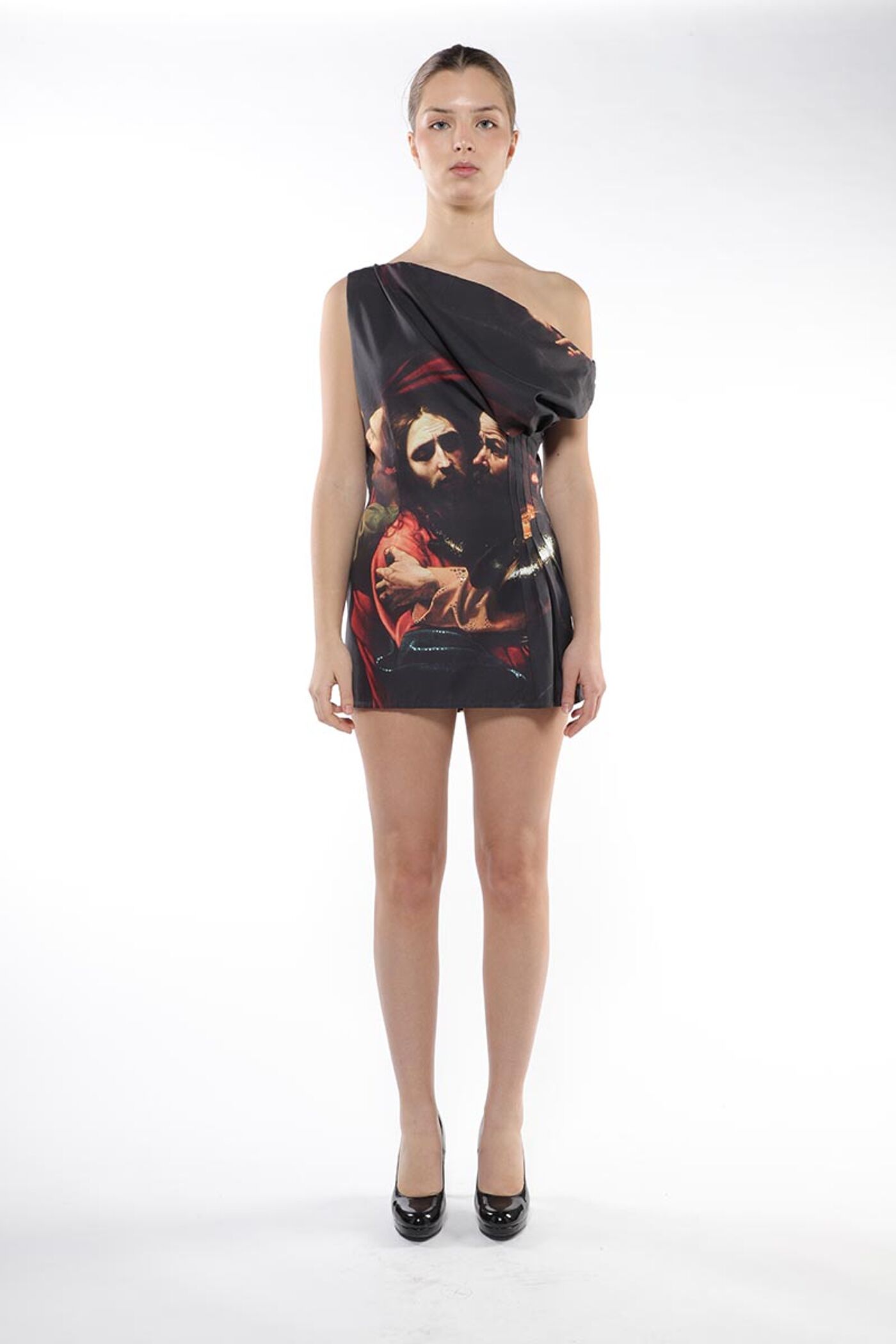 A slender woman with light skin and dark hair, wearing a black one-shoulder dress adorned with a painting of three men embracing, stands against a white backdrop, her black heels visible.