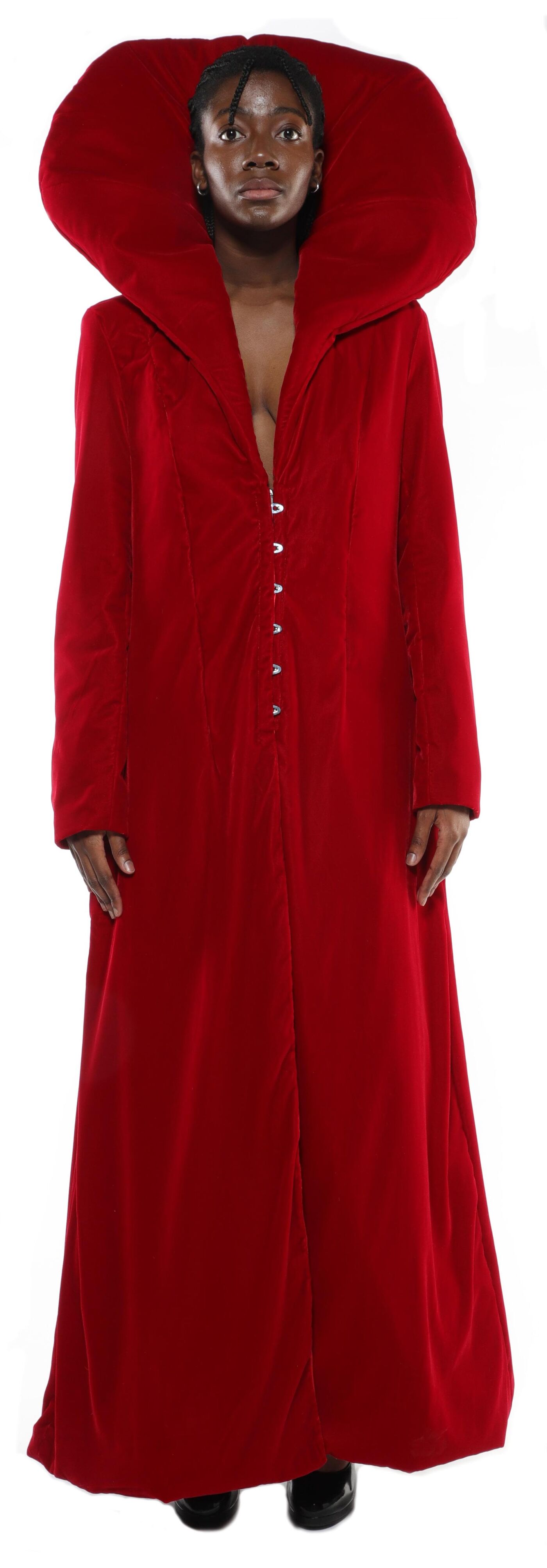A person with dark skin and dark hair is wearing a long, red velvet coat with a large, upturned collar.