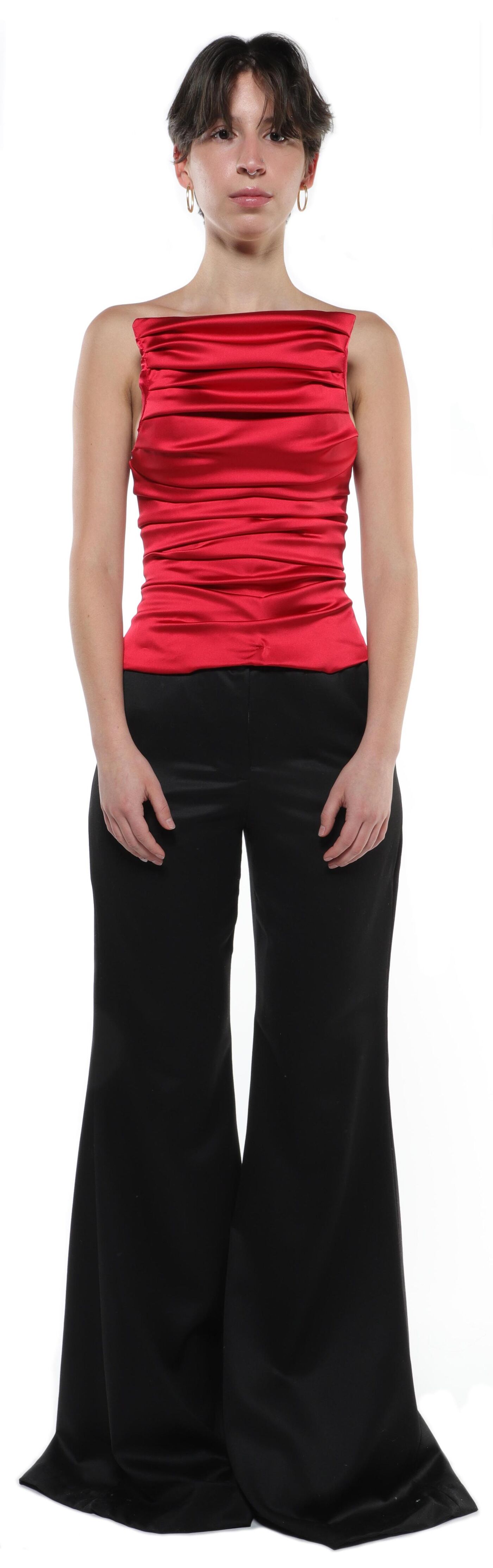 A young woman with short brown hair stands against a white background wearing a red satin cropped top and wide-leg black pants.