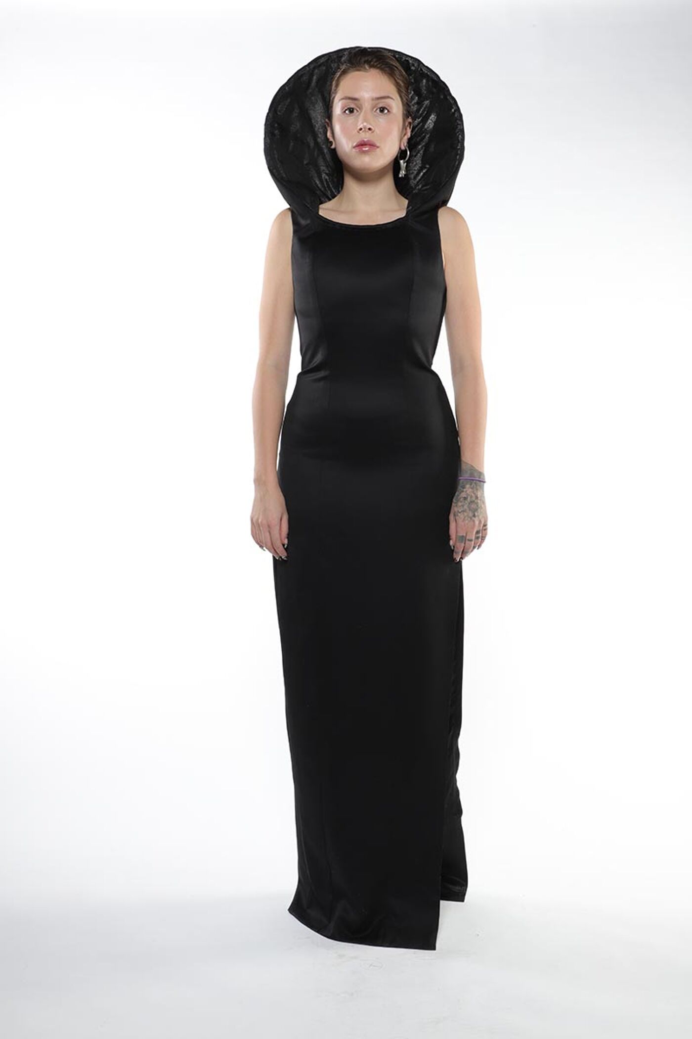 A young woman with a neutral expression stands against a white background, wearing a floor-length black satin dress with a large black fabric collar.