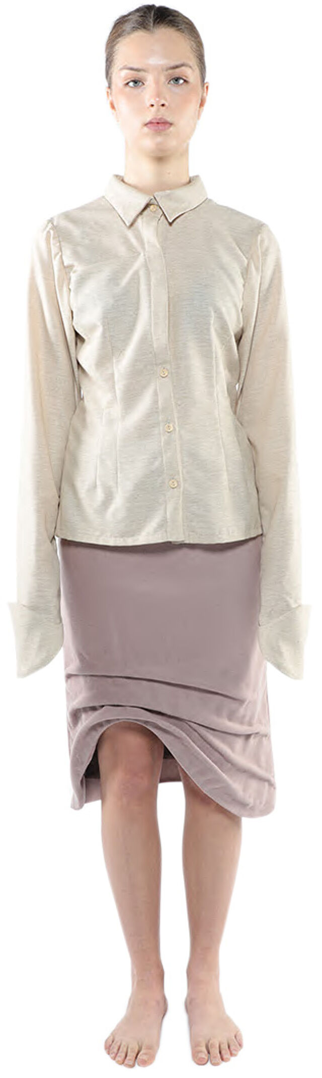 A woman with light skin and dark hair is standing and wearing a long-sleeved, light beige button-down shirt and a mauve, draped skirt.