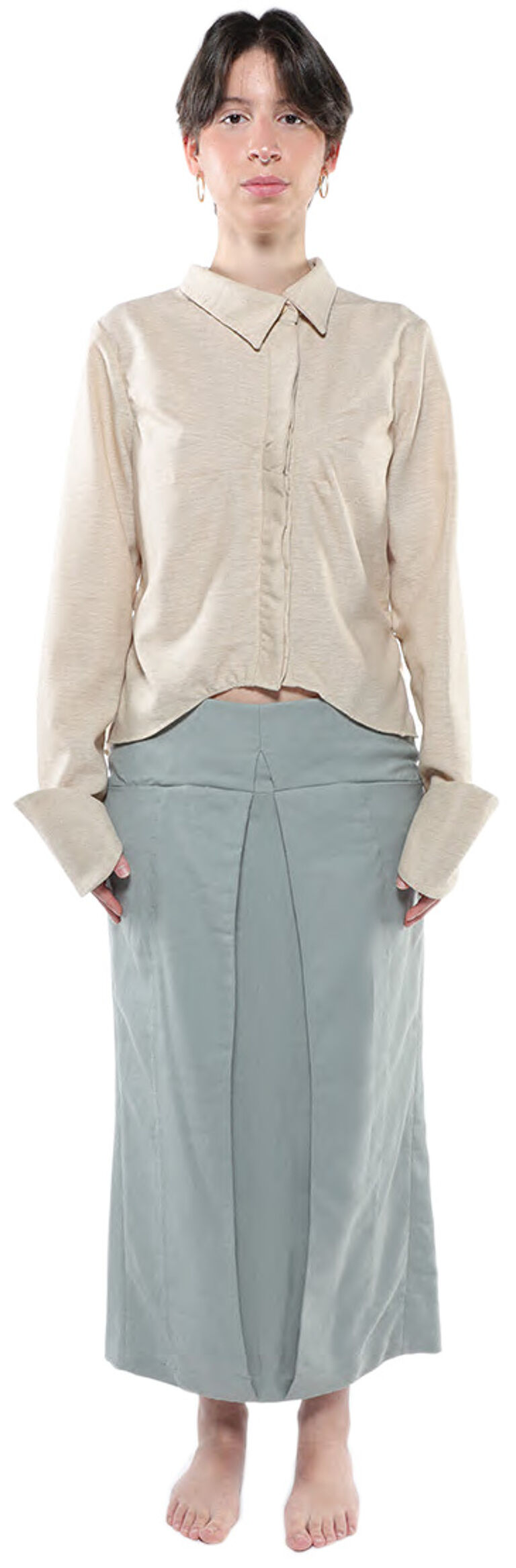 A person with short dark hair is wearing a light beige long-sleeved shirt and a light teal knee-length skirt, standing with their feet slightly apart against a white background.