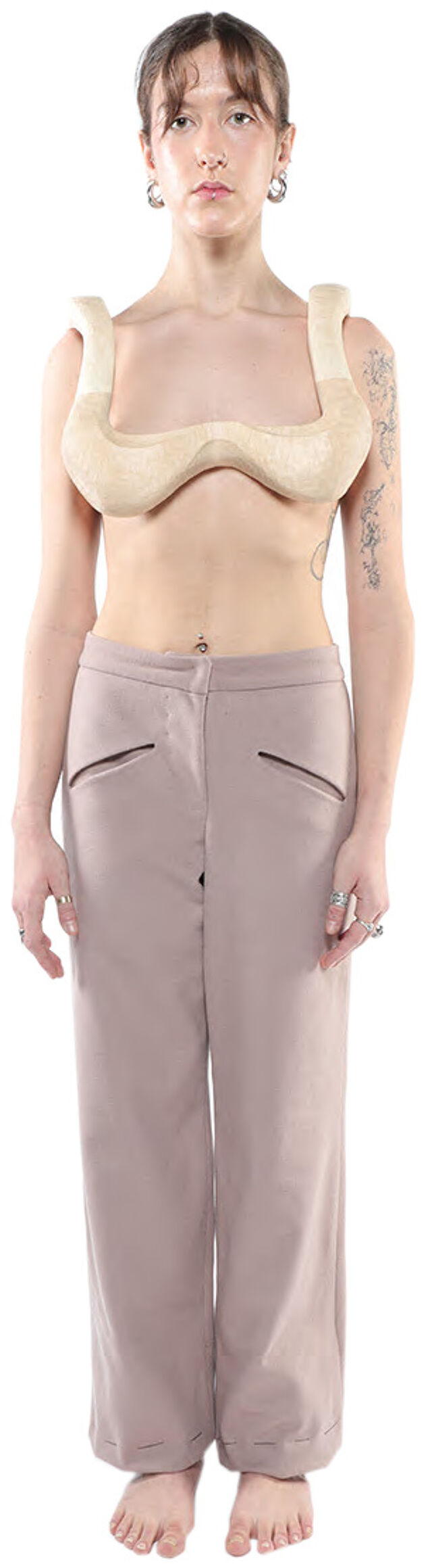A light-skinned woman with brown hair is shown from the knees up wearing a beige, curved, breastplate-like top and loose-fitting, light-brown pants.