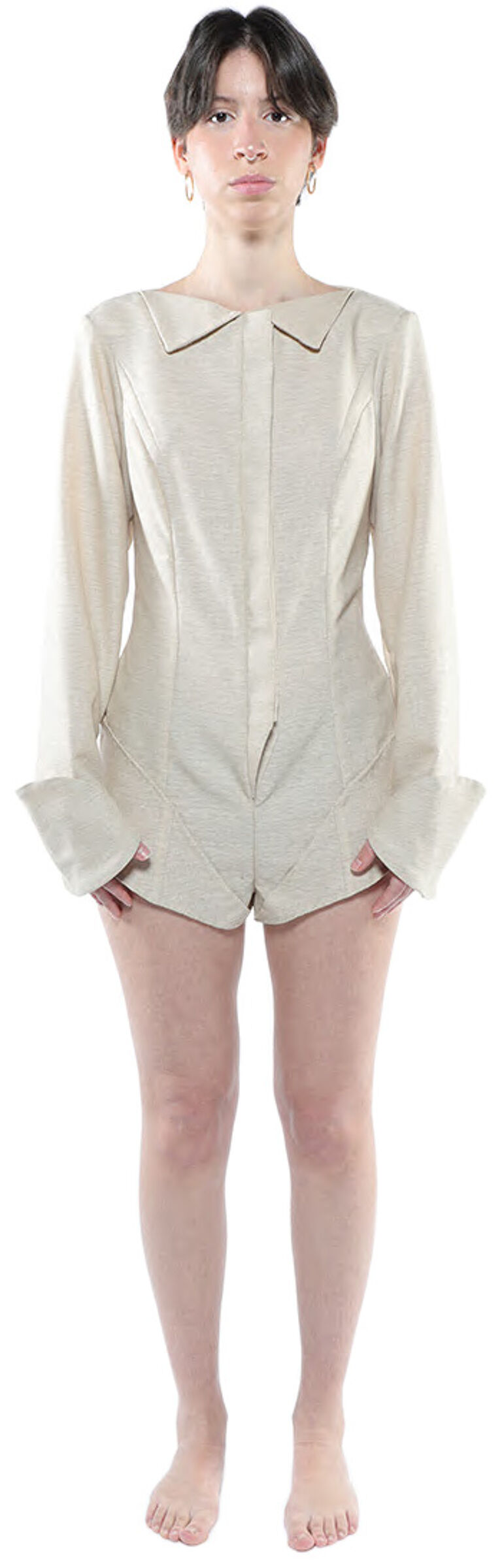 A woman with short dark hair stands against a white background wearing a light beige collared shirt that resembles a jumpsuit.