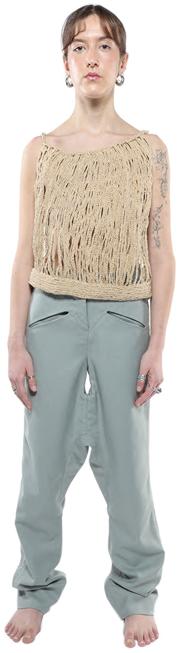 A young woman with shoulder-length brown hair stands against a white background wearing a beige crocheted sleeveless top and loose-fitting light-green pants.