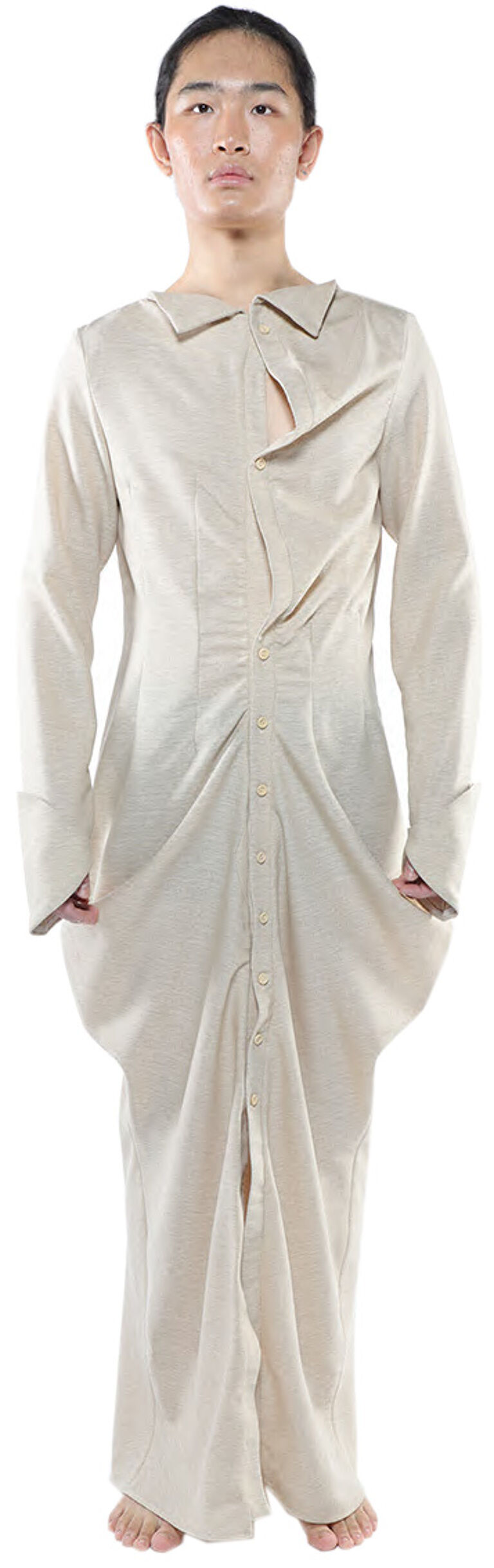 A person with tan skin and dark hair is wearing a long-sleeved, light beige dress with an asymmetrical neckline and a front button closure, standing against a white background.