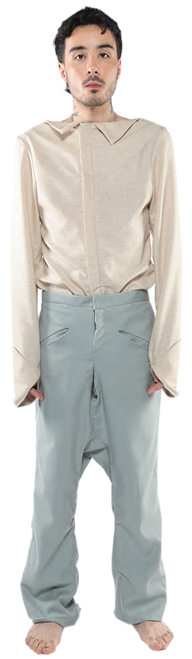 A person with short brown hair is wearing a loose-fitting, long-sleeved beige shirt and light blue pants with a low crotch.
