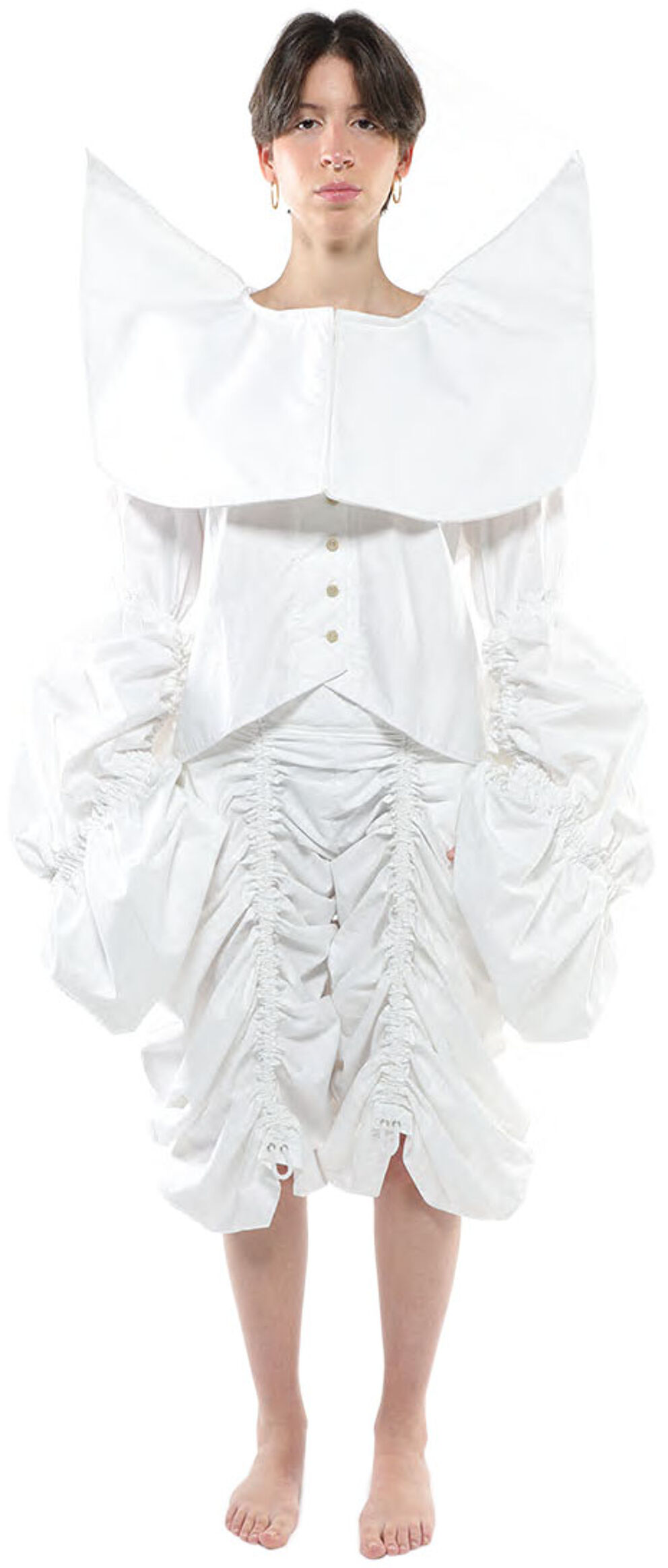 A person with short brown hair wears a white ruffled dress with a large collar and stands with their legs slightly apart against a white background.