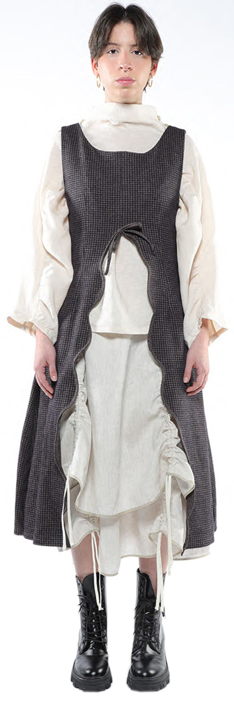 A person with short dark hair is modeling a dark gray, asymmetrical vest over a light beige dress and black combat boots, against a white background.