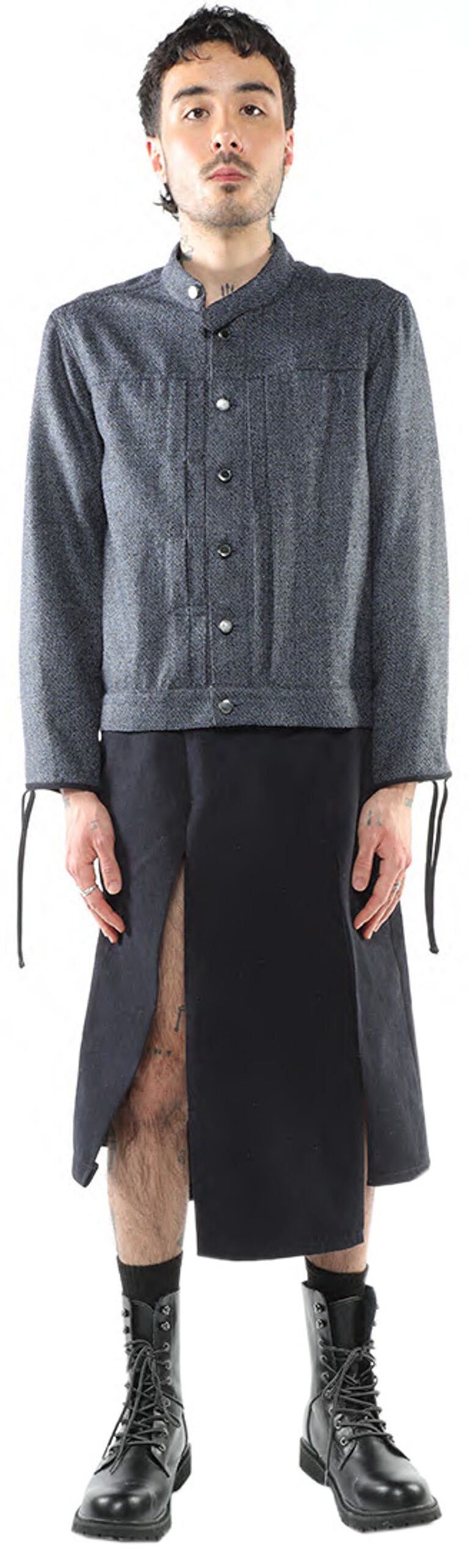 A person with dark hair, wearing a dark gray collared shirt, a black skirt with a slit, and black combat boots, stands against a white background.