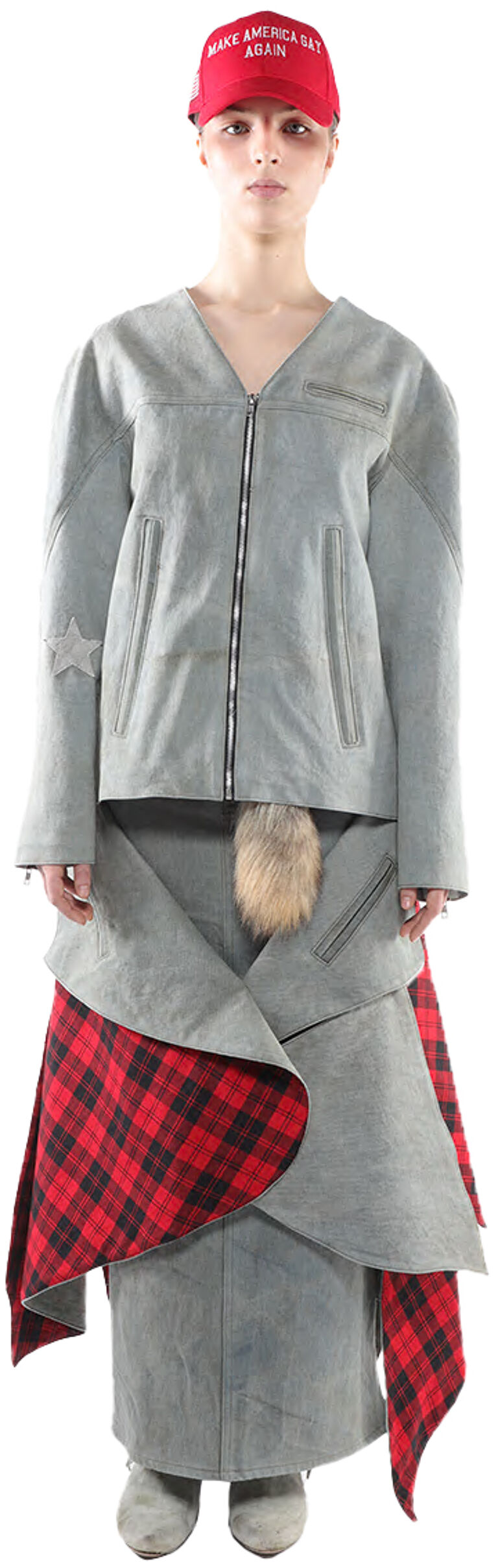 A person wearing a gray jacket and skirt, a red and black plaid wrap, and a red "Make America Great Again" hat stands against a white background.