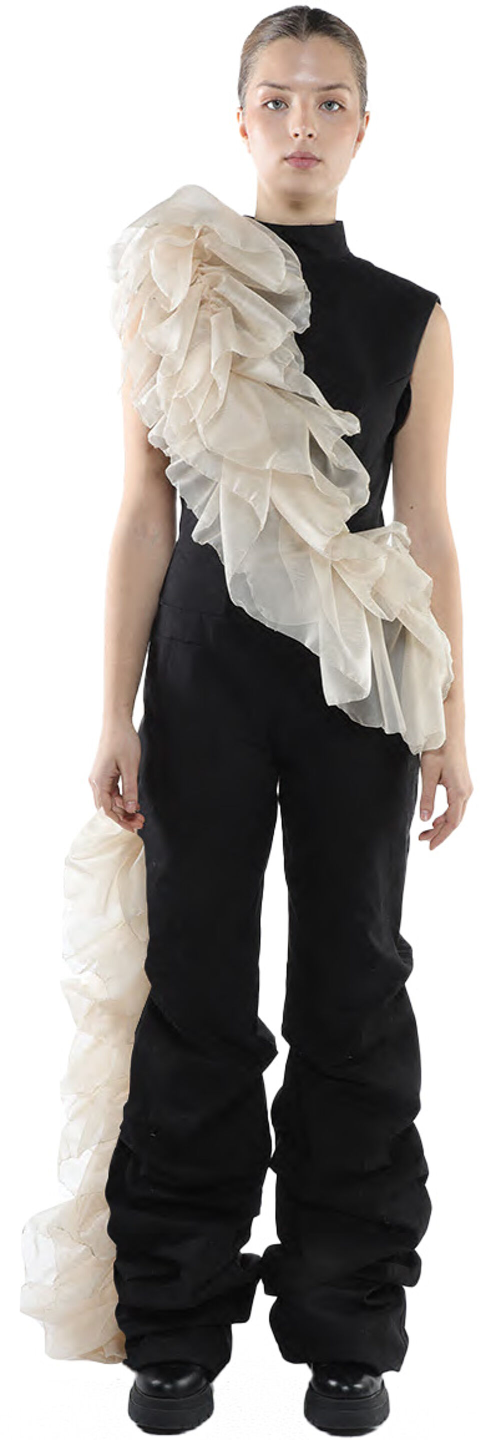 A woman with light skin and dark hair is modeling a black jumpsuit with a large, off-white ruffled overlay on one shoulder, and the pants are gathered at the ankles.