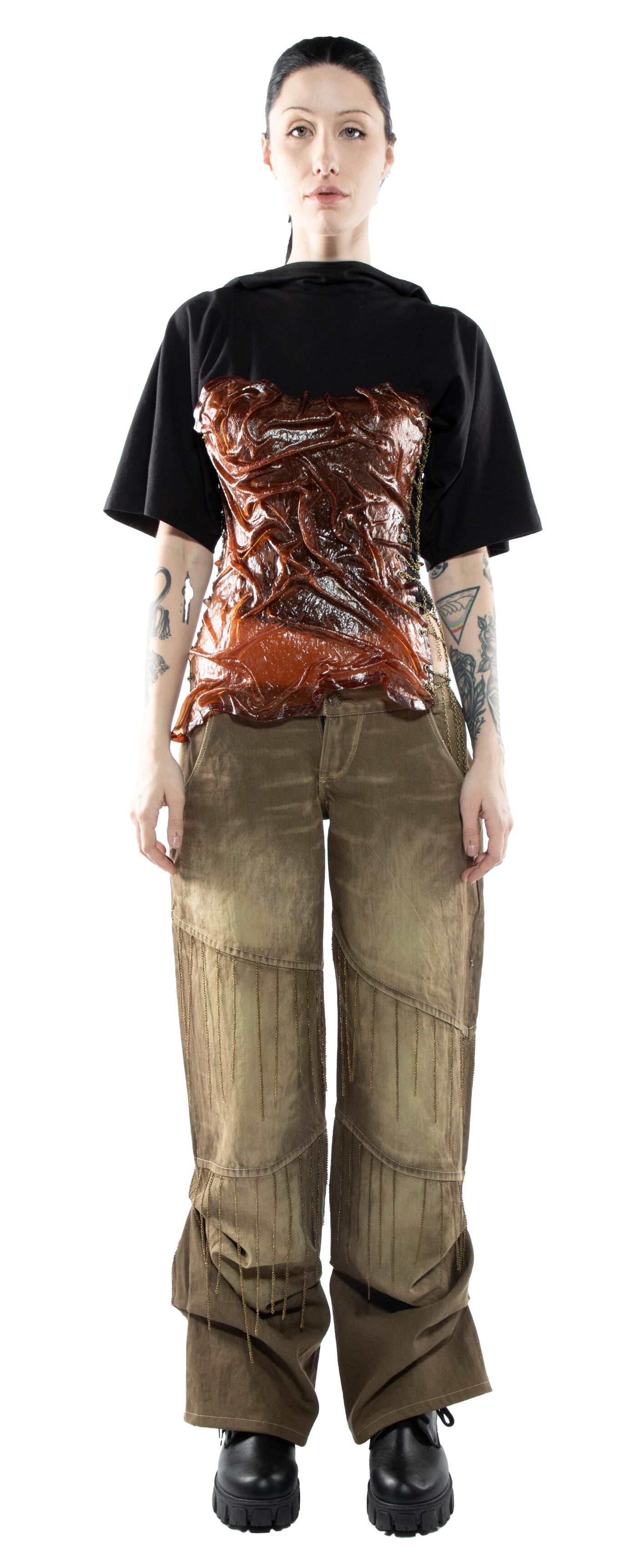 A person with dark hair and tattoos is wearing a black short-sleeved shirt over a brown corset and loose-fitting, light-brown pants with dark shoes.