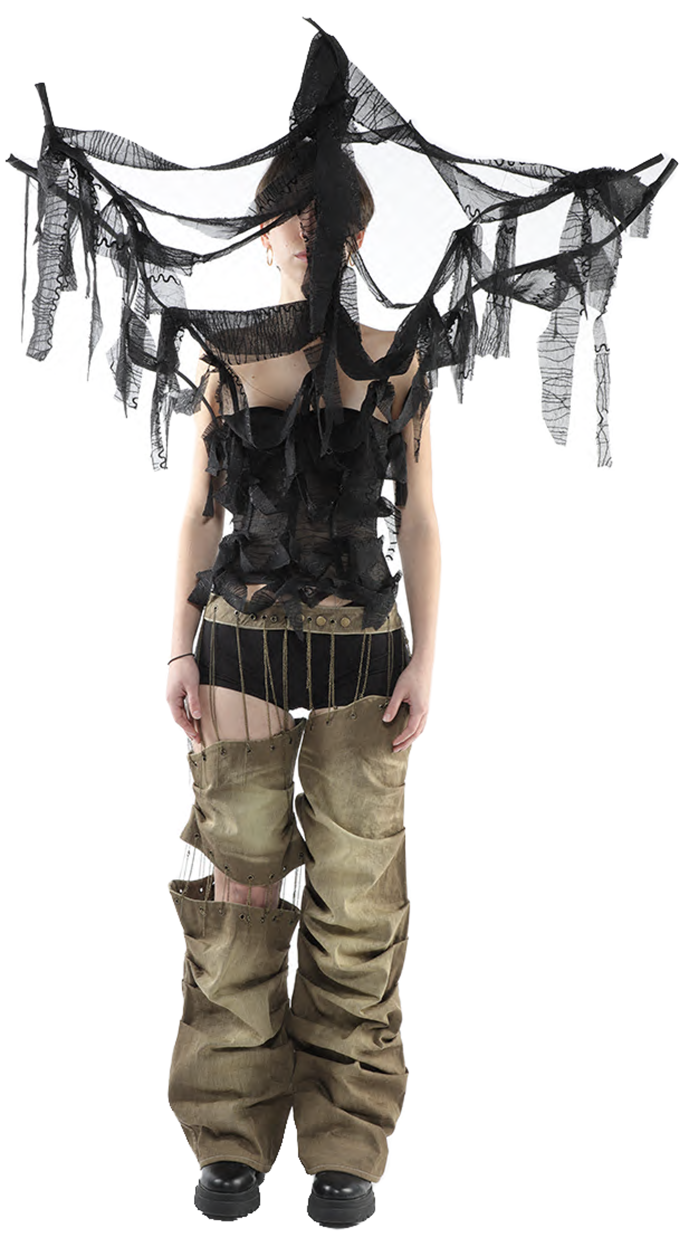 A person with short dark hair wears a tattered black fabric headpiece and top over olive green pants with cut-out knees and dark shoes, standing against a white background.