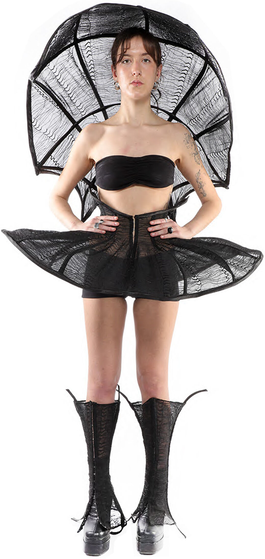 A model wears a black, lacy, wide-brimmed hat and matching short dress and tall boots, posing against a white background.