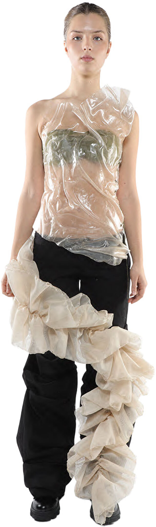A model with fair skin and dark hair wears a sheer, one-shoulder top over black pants, adorned with a large, ruffled beige fabric piece cascading down one leg.