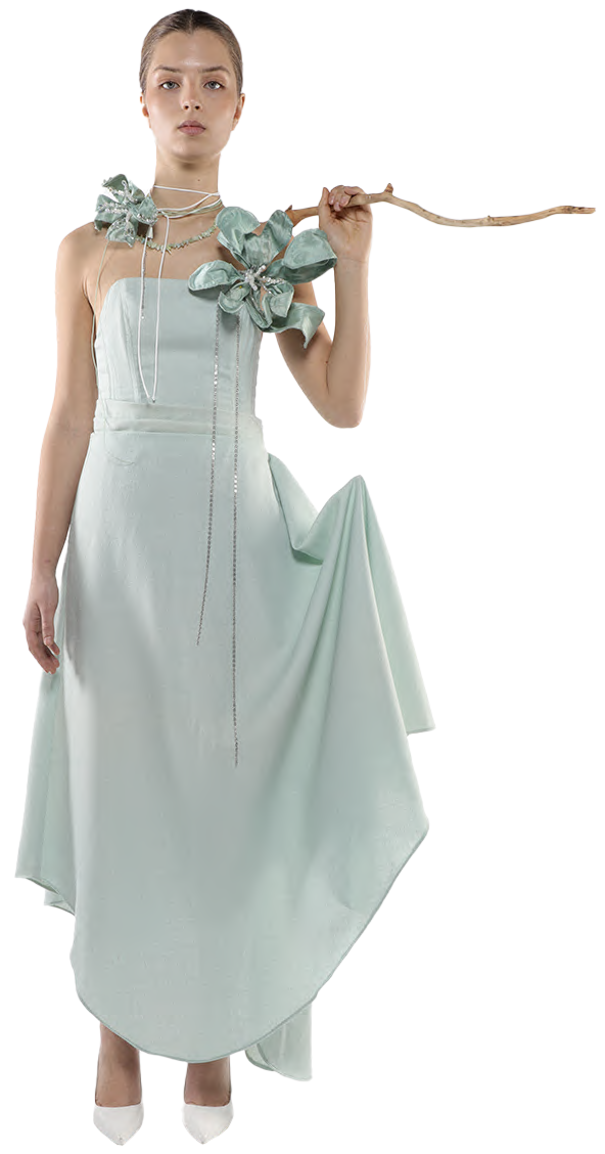 A slender woman with light skin and dark hair, wearing a pale green, asymmetrical gown adorned with light green fabric flowers and a long, delicate necklace, holds a slender branch across her shoulders.