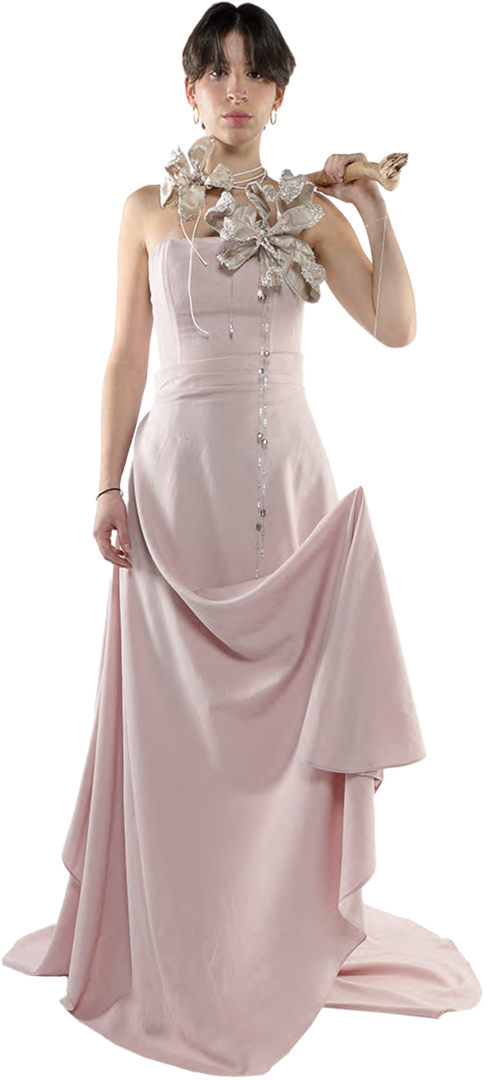 A young woman with short dark hair wears a long, light-pink gown with a unique embellished shoulder piece and holds a light-brown, possibly wooden, object across her shoulder.