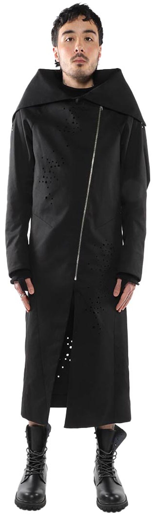 A man wearing a black, long-sleeved coat with cutouts and lace-up boots stands against a white background.
