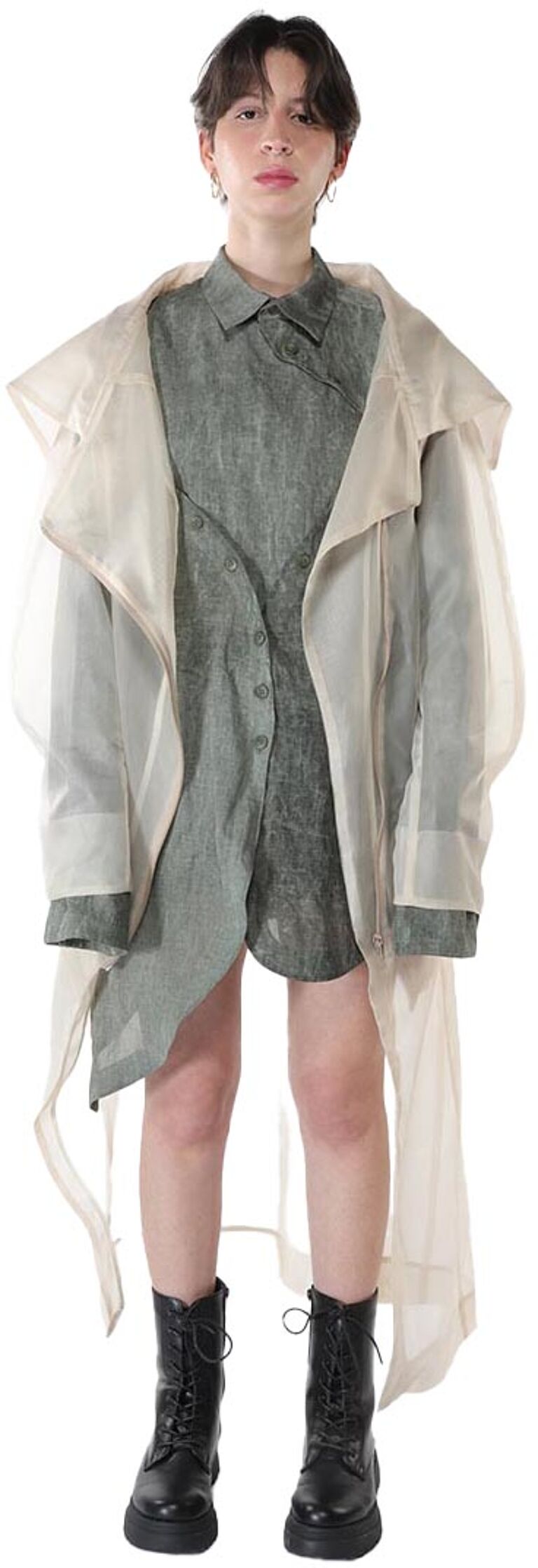 A person with short dark hair is wearing a long, sheer beige coat over a dark green button-down shirt and black combat boots.