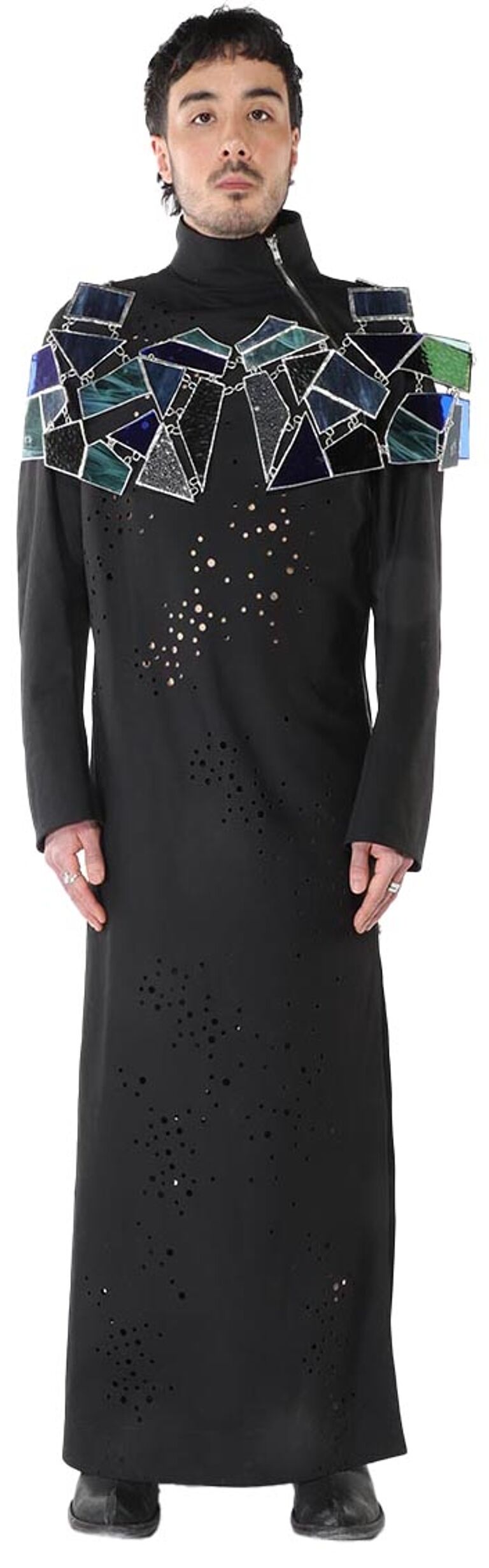 A person with short brown hair is wearing a long black dress with a colorful stained-glass-like pattern on the shoulders and laser-cut detailing on the body.