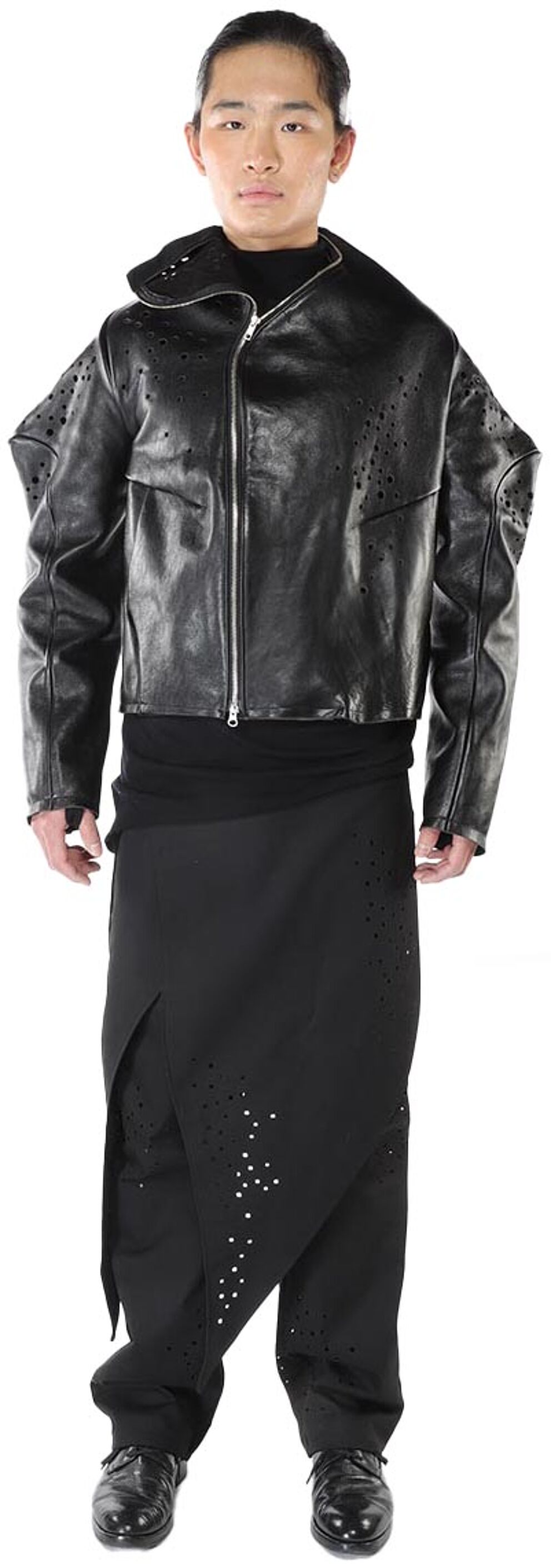 A person with dark hair and skin is modeling a black leather jacket with cut-out detailing and matching black, asymmetrical pants with similar cut-outs, standing against a white background.