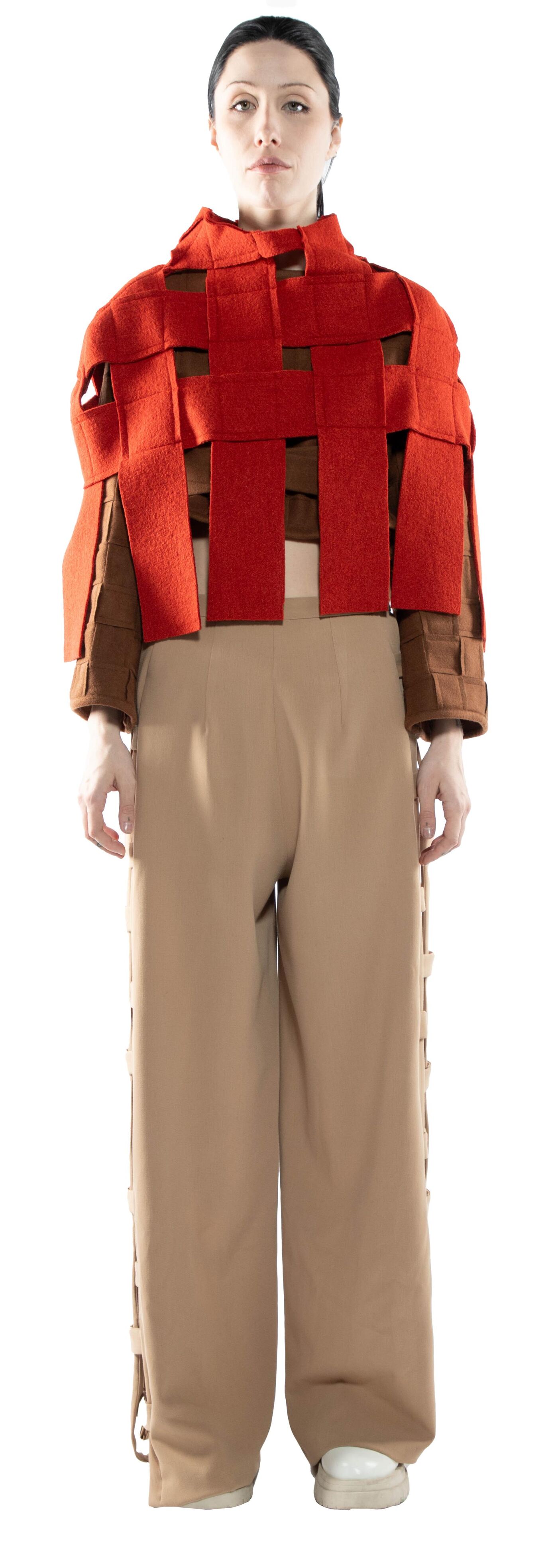 A woman with dark hair is standing and wearing a red woven top and light brown pants.
