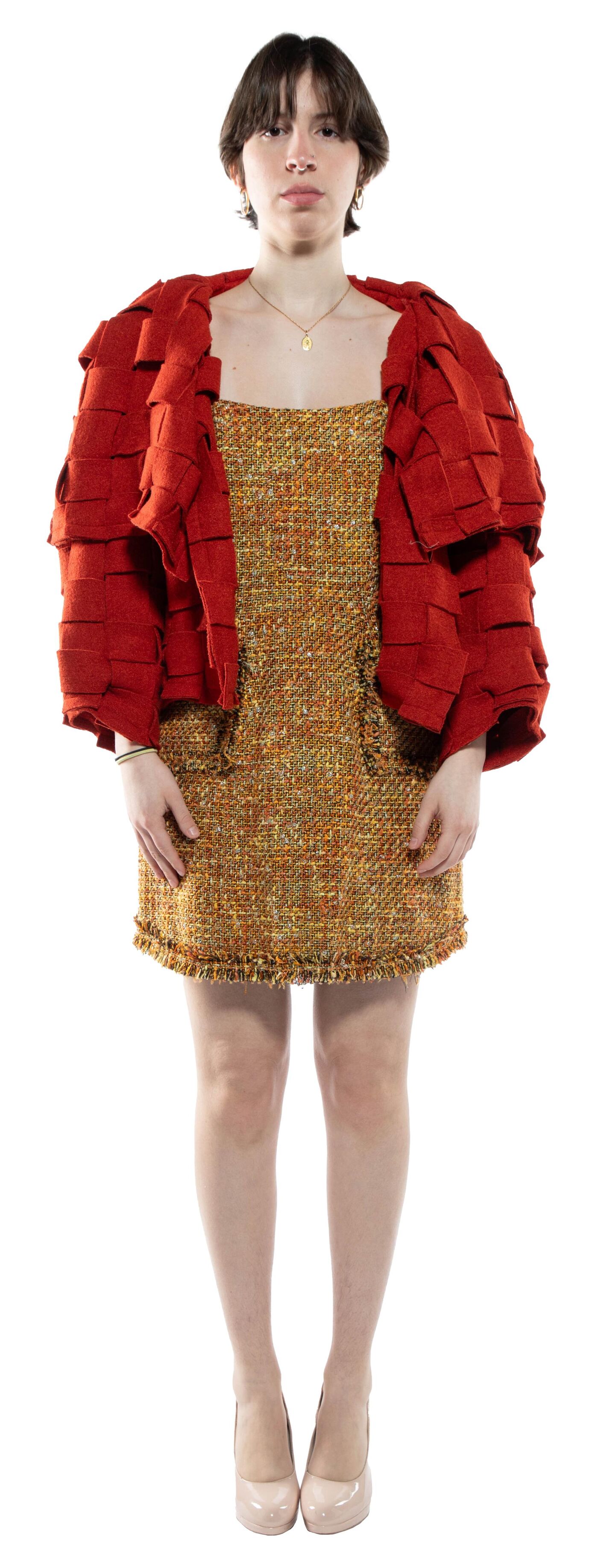 A woman with short dark hair, wearing a rust-colored layered jacket over a gold tweed mini dress and nude heels, stands against a white background.