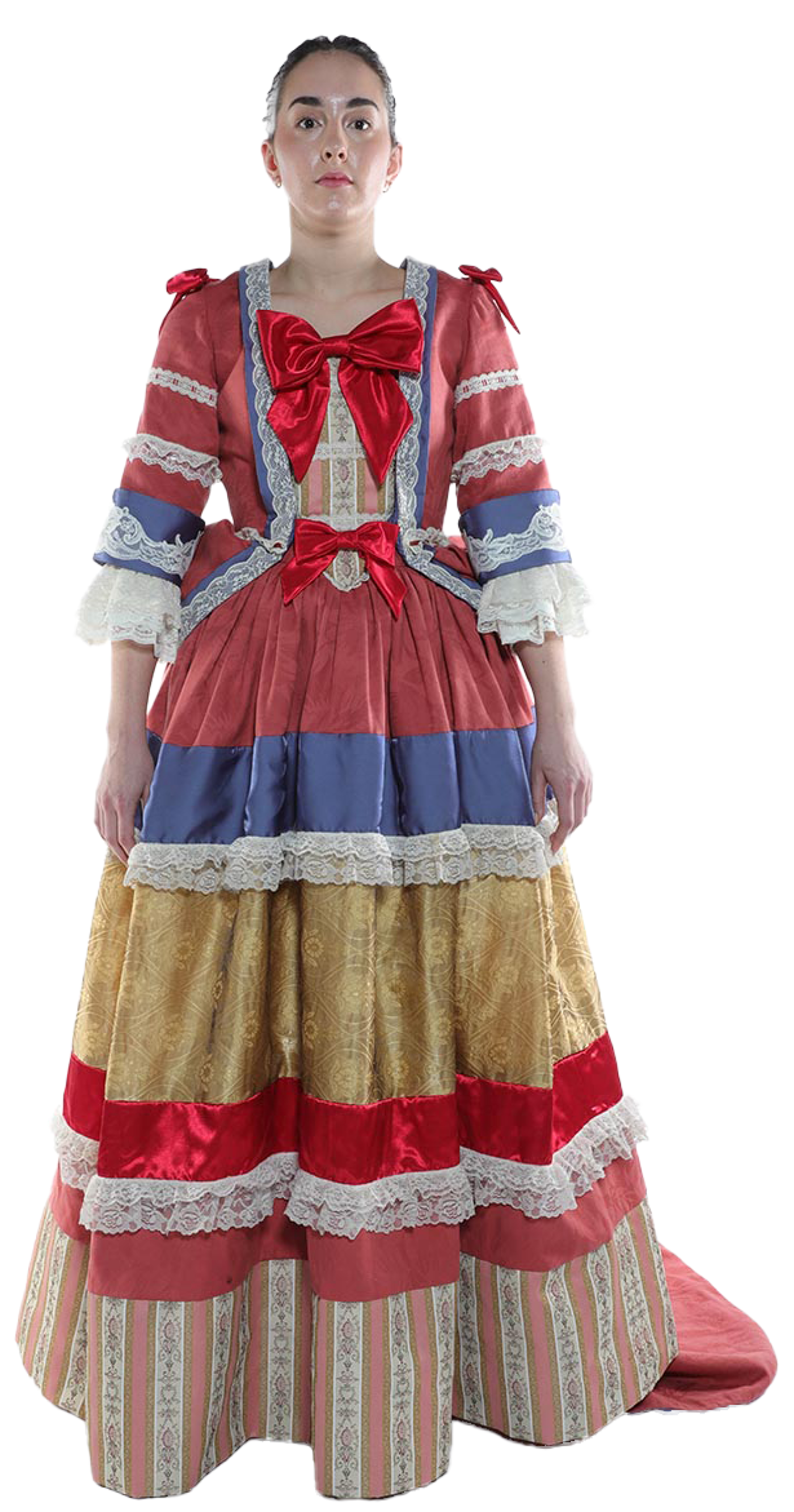 A woman stands against a white background, wearing a long, multi-layered gown with red, gold, blue, and pink sections, accented with lace and ribbons.