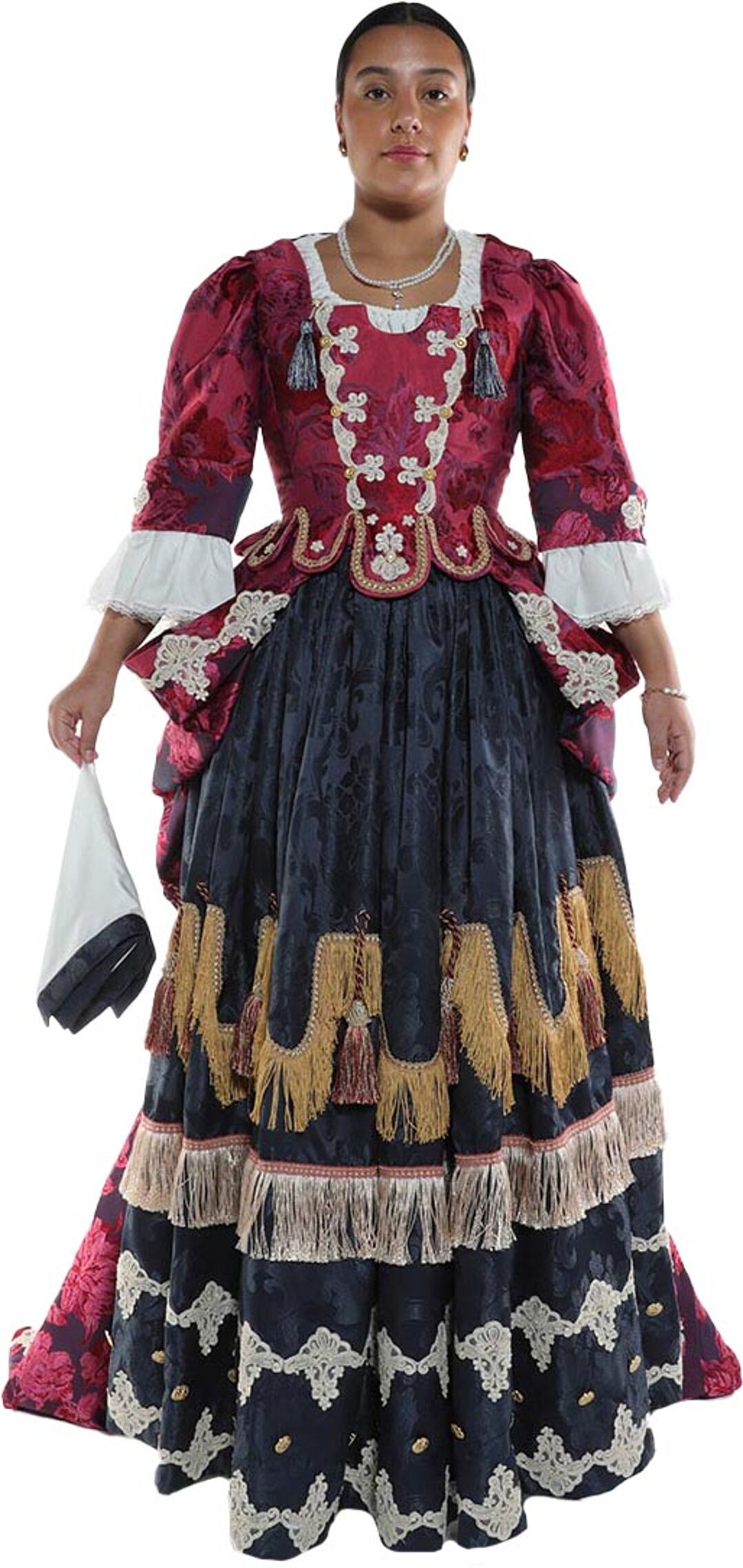 A woman with dark hair and light skin is modeling a long, richly decorated burgundy and black gown with gold and white trim, holding a white handkerchief.