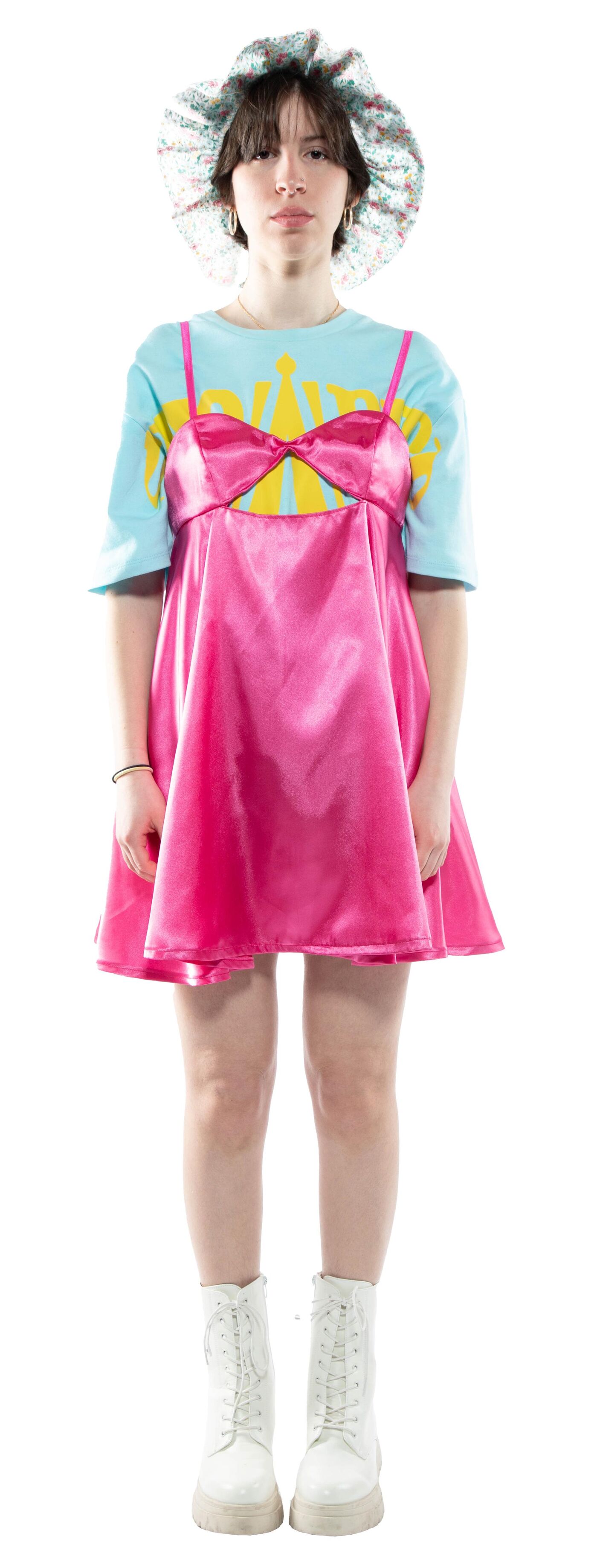 A young woman with short brown hair wears a pink satin slip dress over a light blue t-shirt, a floral print hat, and white combat boots.