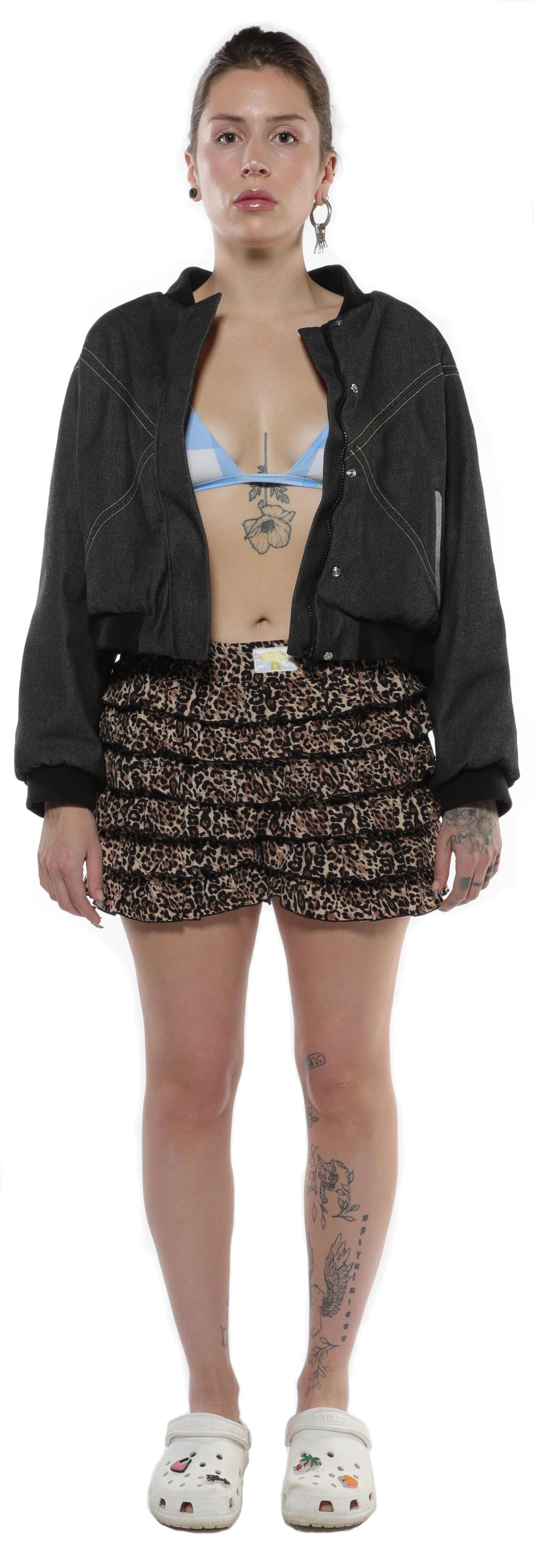 A young woman with tattoos on her legs wears a dark gray cropped bomber jacket over a light blue bralette and leopard-print ruffled shorts, paired with white clogs.