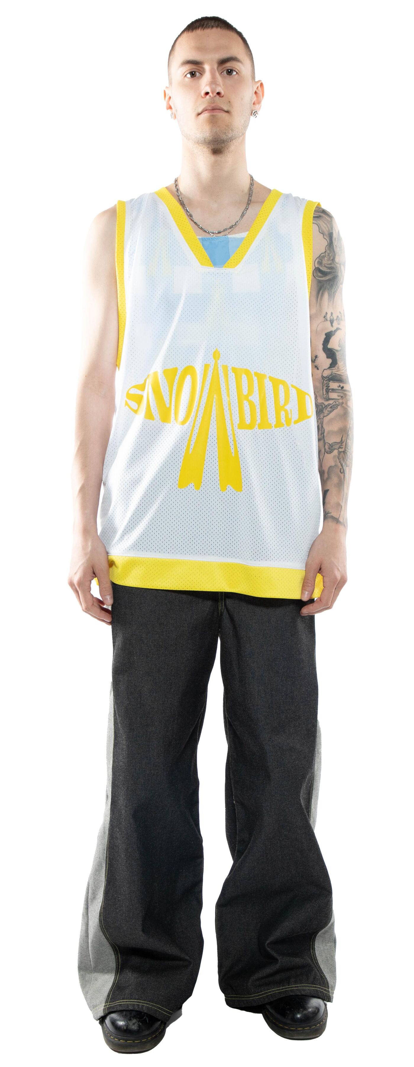 A light-skinned man with tattoos on his arms models a white basketball jersey with yellow lettering and dark grey flared pants against a white background.