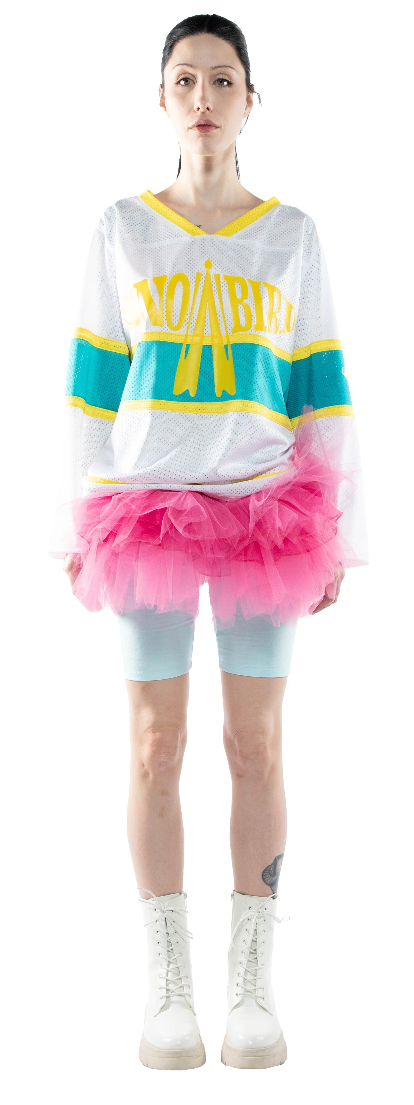 A woman with dark hair wears a white jersey with teal and yellow accents, a pink tulle skirt, light blue shorts, and white combat boots.