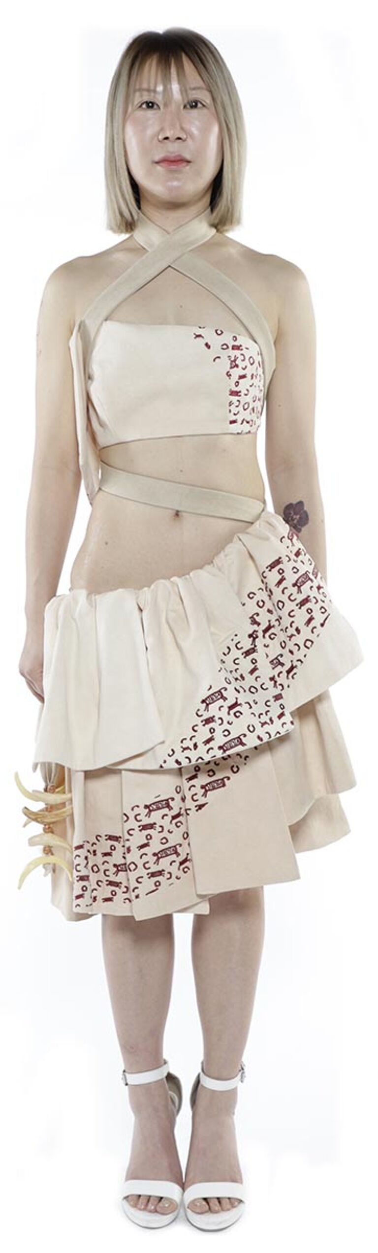 A light-skinned woman with short blonde hair models a beige asymmetrical dress with a pattern and white heels.