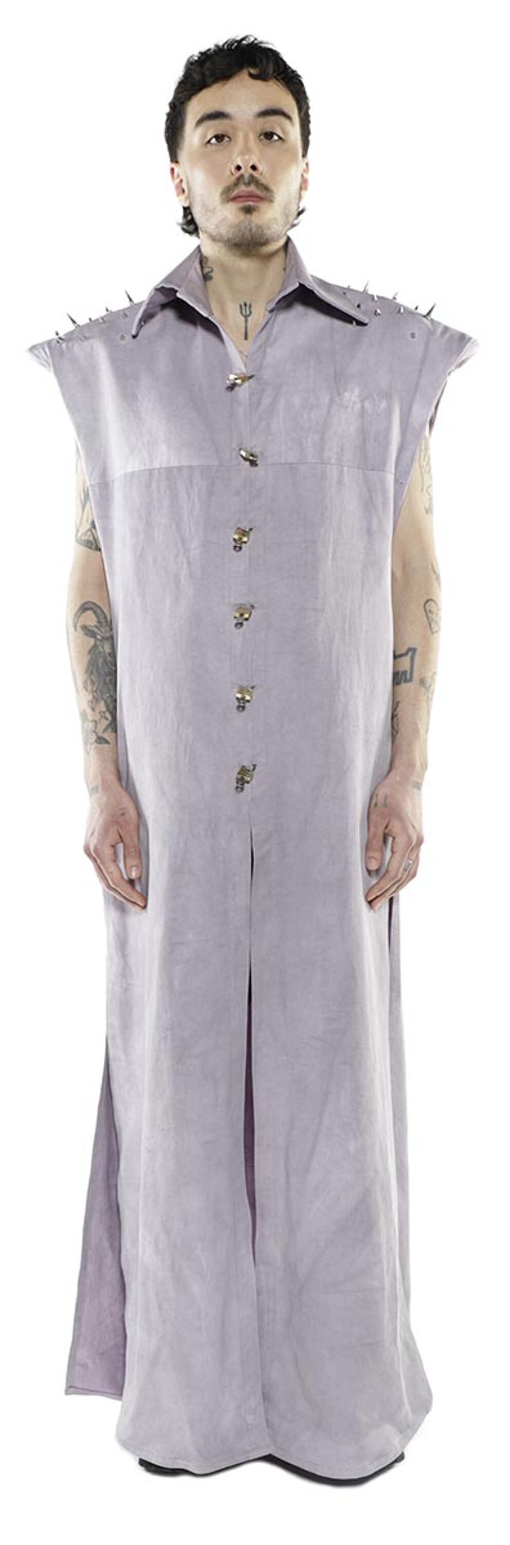 A person with tattoos stands against a white background wearing a long, sleeveless, light purple collared shirt with studded shoulders and metal buttons.