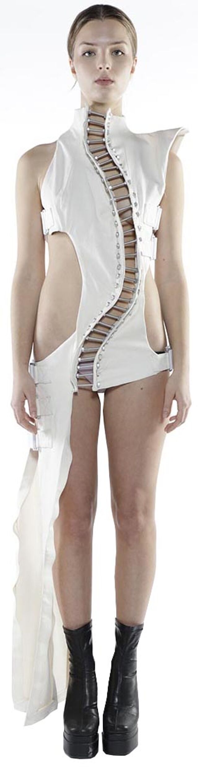 A slender woman with light skin and dark hair, wearing a white asymmetrical dress with a unique beaded detail and black platform boots, stands against a stark white background.