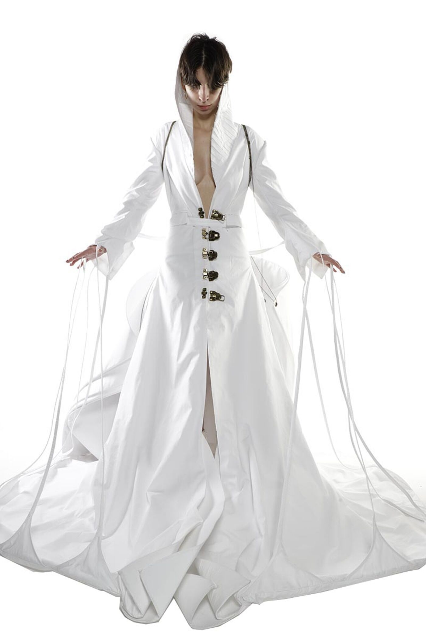 A woman with short dark hair wears a long white coat with a hood and buttons down the front, standing against a white background with long white fabric trailing from the sleeves and bottom of the coat.