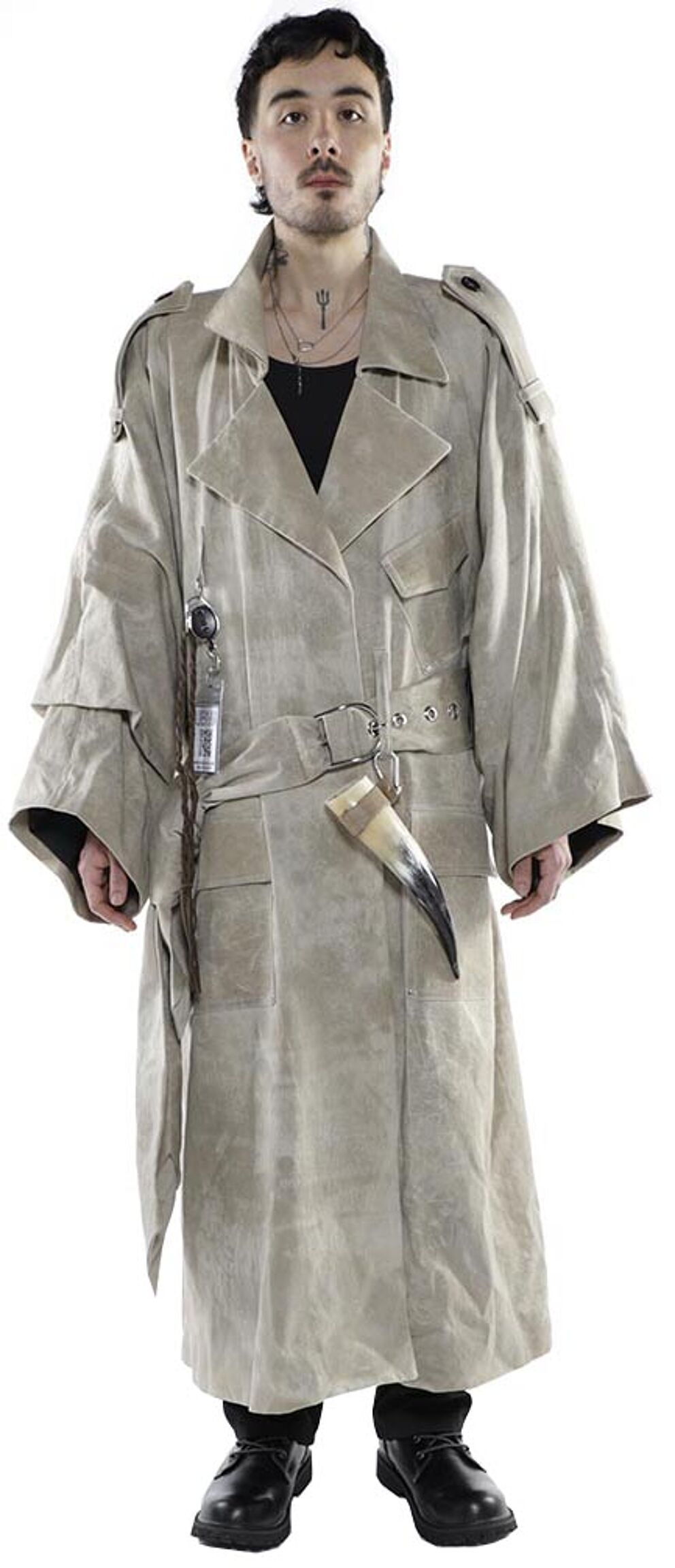 A person with dark hair and a pale complexion wears an oversized, light beige trench coat with various accessories hanging from it, standing against a white background.