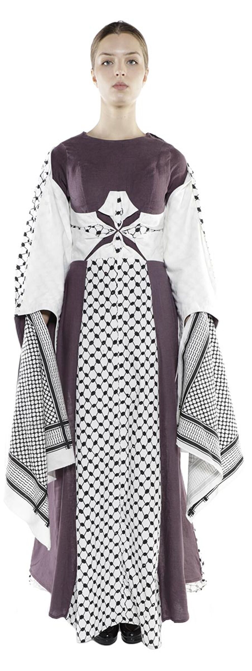 A woman is standing and wearing a long dress with a brown bodice and white and black checkered keffiyeh sleeves.
