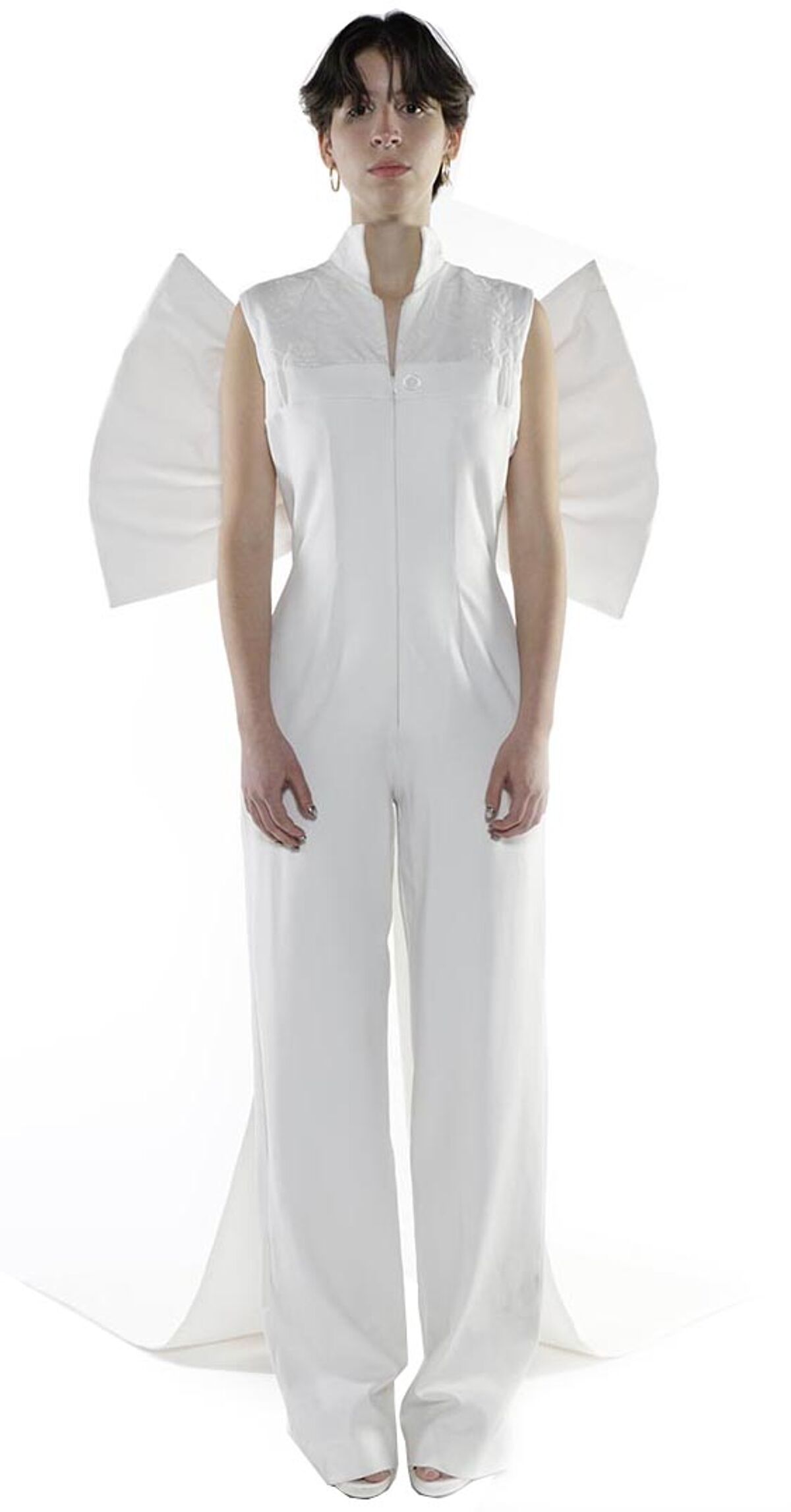 A woman with short dark hair wears a white jumpsuit with large, wing-like fabric pieces attached at the shoulders.