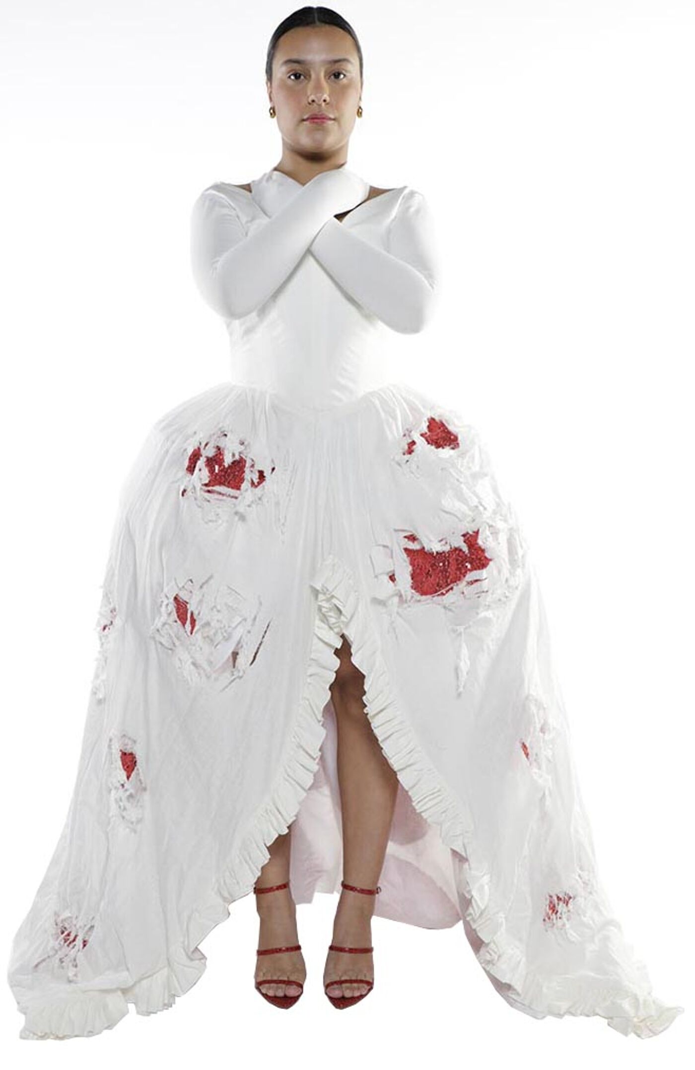 A young woman with crossed arms wears a long, white, tattered dress with red accents and red high-heeled shoes.