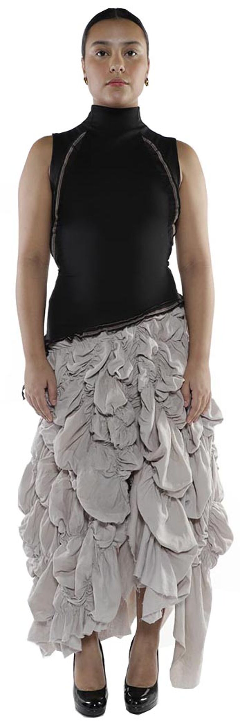 A woman with tan skin and dark hair is wearing a black turtleneck sleeveless top and a light-grey ruffled maxi skirt, and black heels.